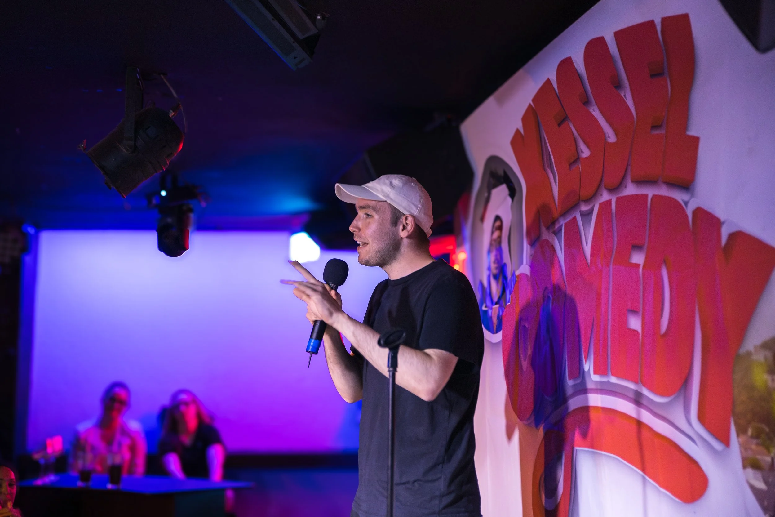 Ein junger Mann trägt eine weiße Cap, hält ein Mikrofon und spricht auf einer Bühne bei einem Comedy-Event, im Hintergrund sitzen zwei Frauen, die zusehen, mit einem beleuchteten Bildschirm und einem Banner mit der Aufschrift 'Nedell Comedy'.