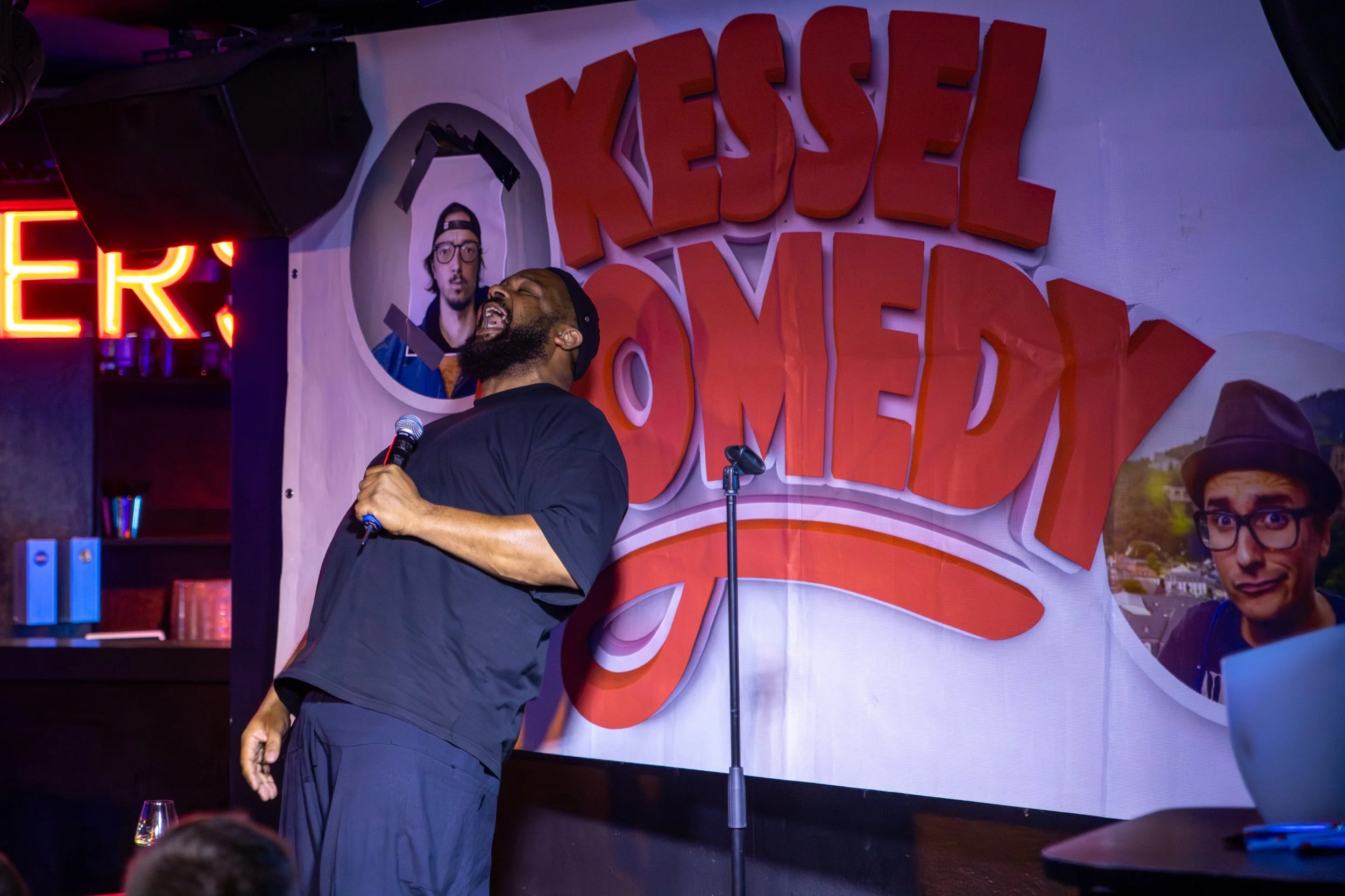 Ein Mann mit dunklem Bart singt lachend auf einer Bühne bei einer Comedy-Show. Im Hintergrund ist ein Banner mit der Aufschrift 'Kessel Comedy' und einem Profilfoto eines anderen Mannes. Es gibt eine Mikrofonstange, einen Laptop auf einem Tisch und e