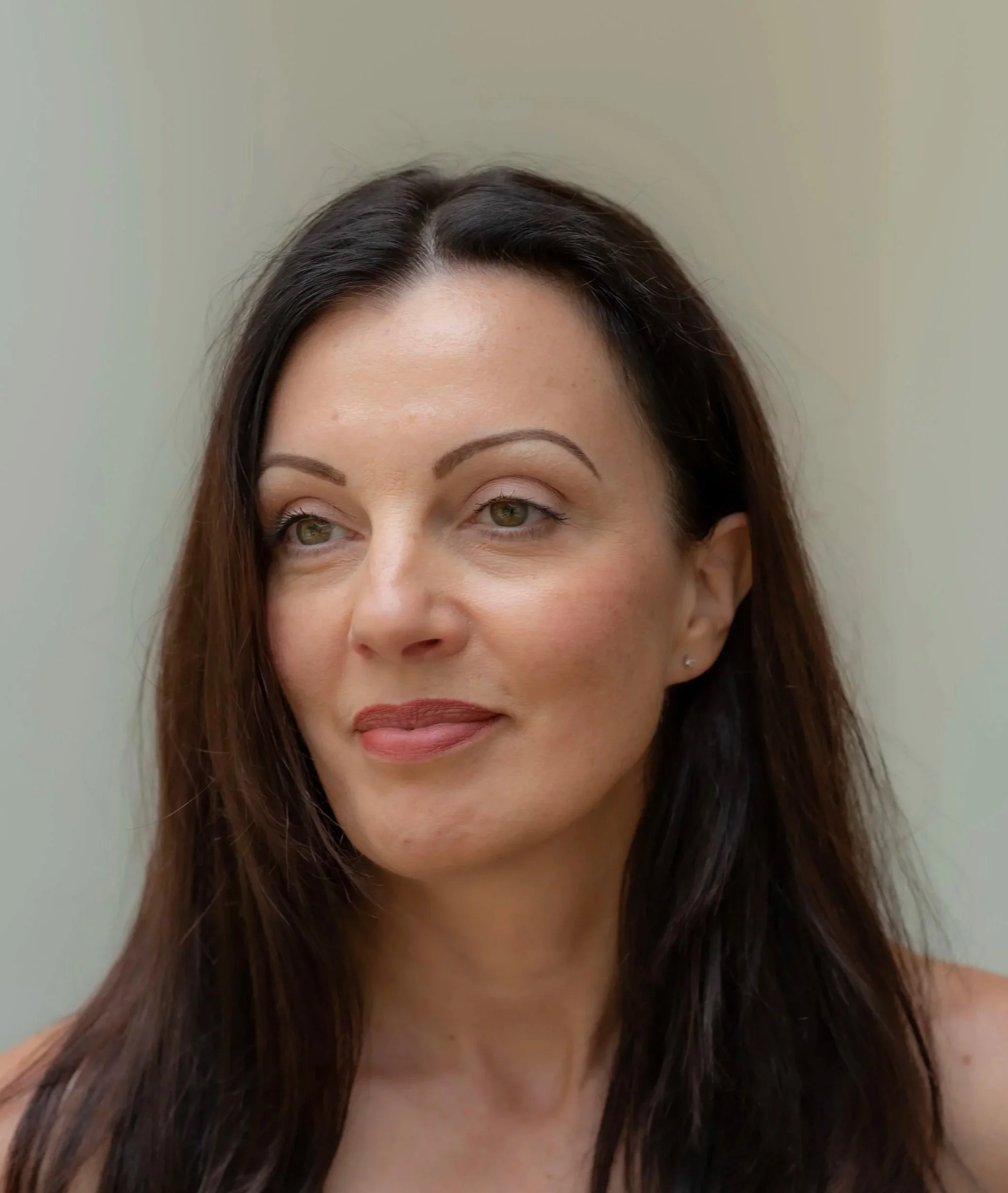 Close-up of a woman's face, showing her forehead, eyes, eyebrows, nose, and part of her mouth and chin. She has long brown hair, light makeup, and a neutral expression. The background is plain and light-colored.