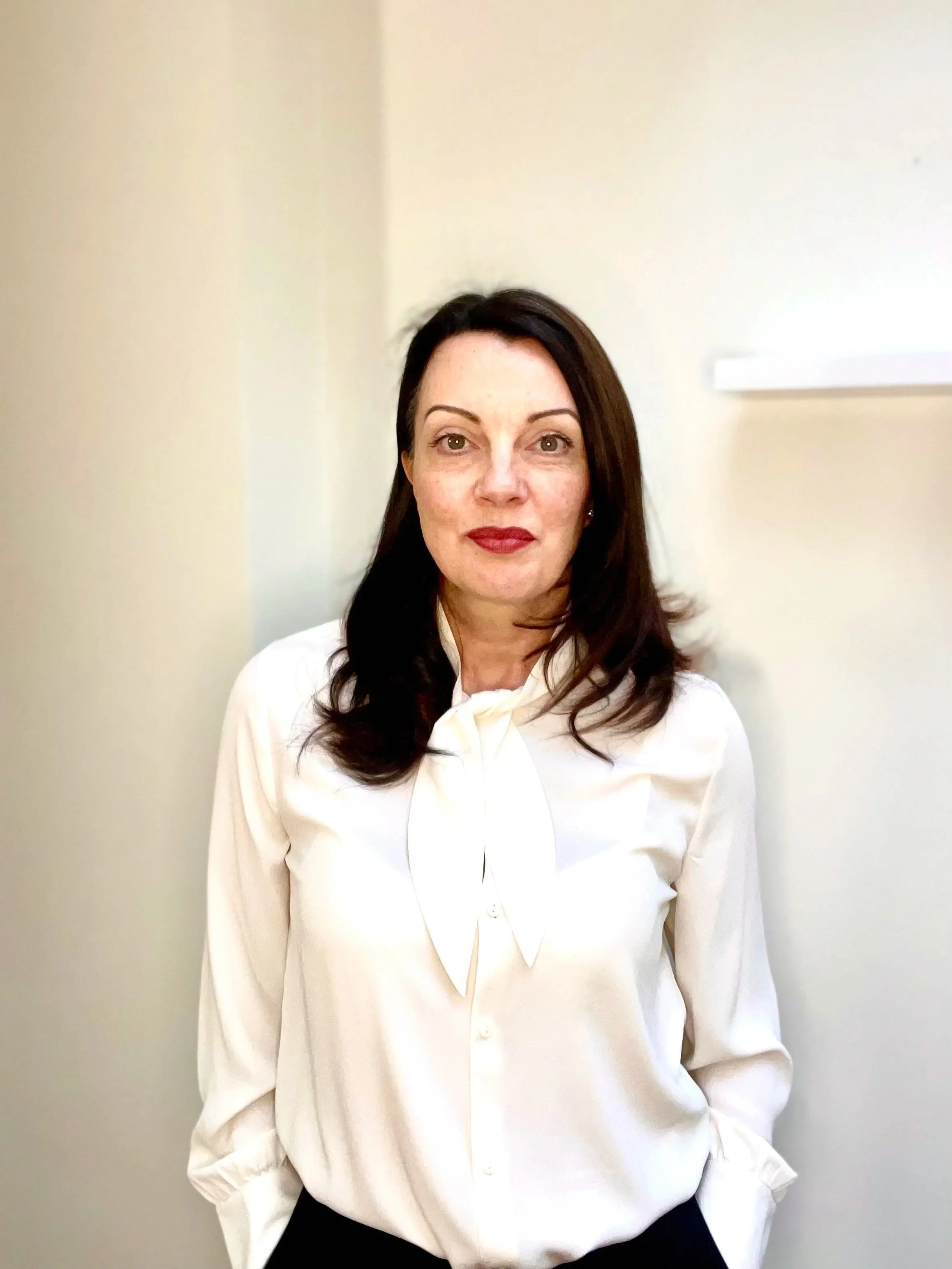 A woman with dark hair, wearing a white blouse with a bow, standing against a plain off-white wall, looking directly at the camera.