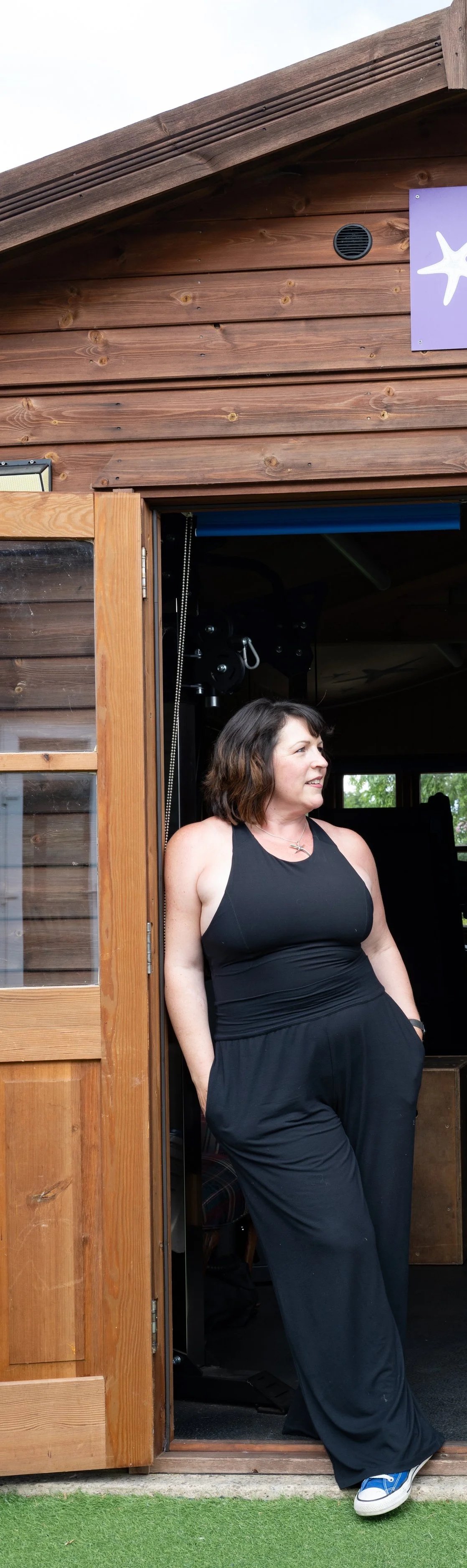 Founder & co-director Sara Southey, a woman with shoulder-length brown hair wearing a black tank top, black pants, and white sneakers standing at a wooden door of the shed of strength, with a starfish sign on the exterior wall.