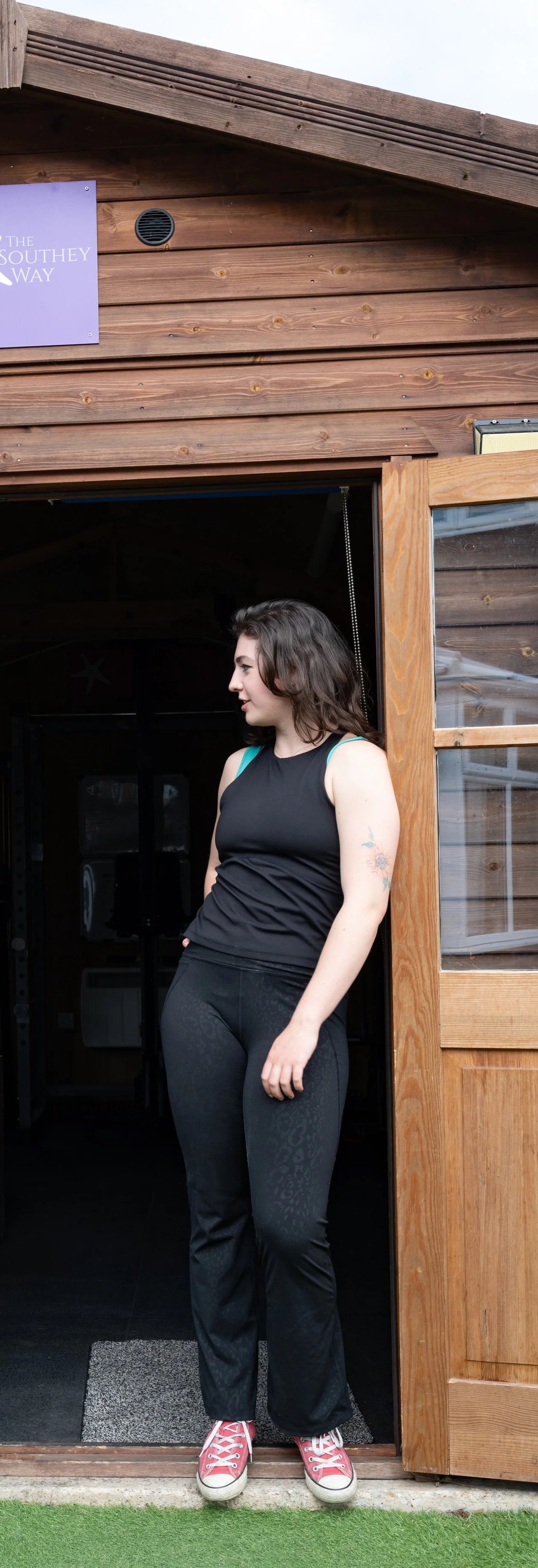 Co-director Jenny Southey, a woman with wavy brown hair standing at a wooden door of the shed of strength, wearing a black sleeveless top with teal straps, black pants with a subtle pattern, and red sneakers. She has a tattoo on her left arm.