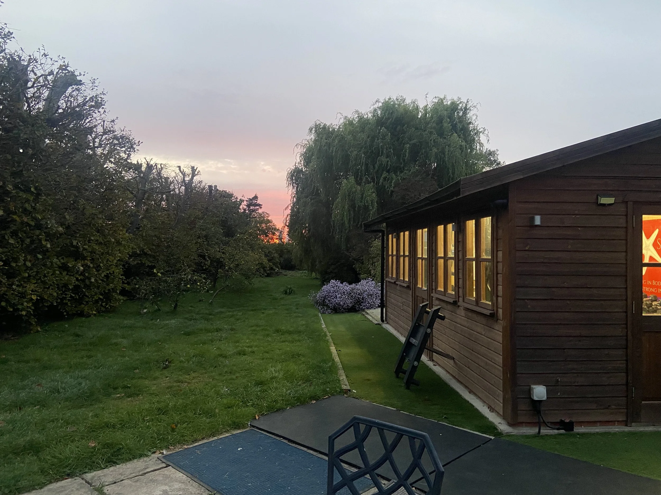 Evening view of a garden with a wooden building called the shed of strength, illuminated windows, and a sunrise sky in the background.
