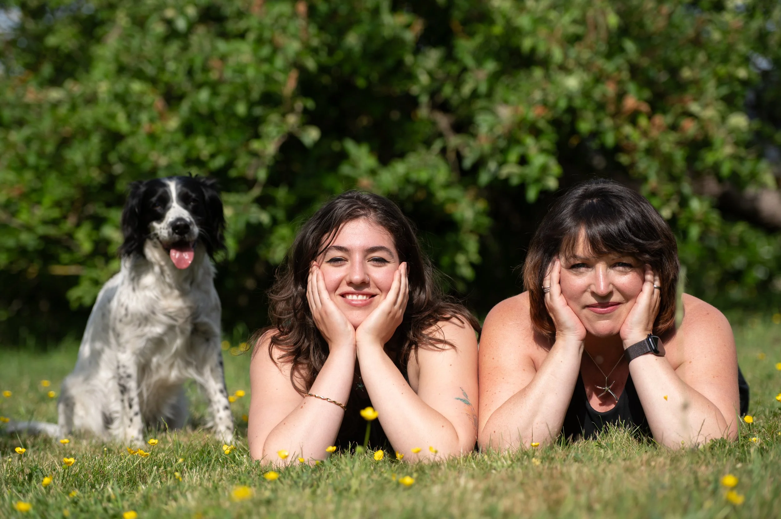 Co-directors Sara & Jenny and a dog lying on grass with yellow flowers, smiling, with green trees in the background.
