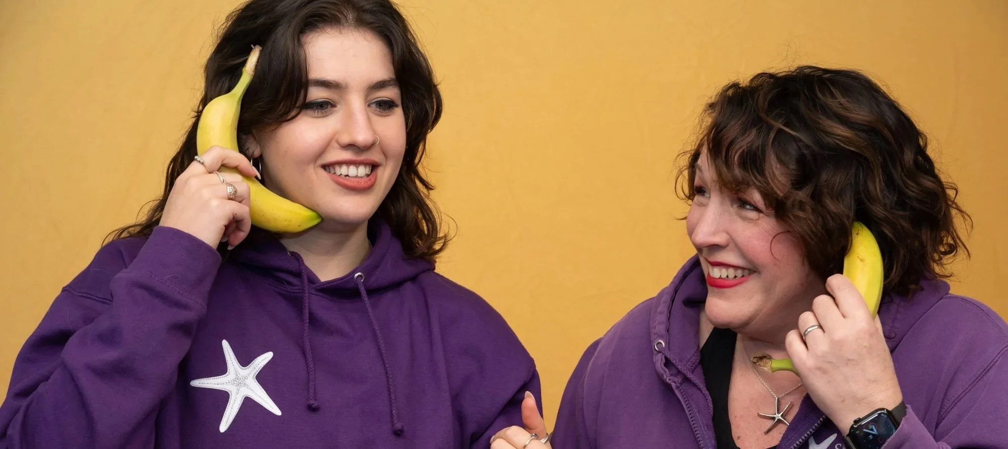 Co-directors Sara & Jenny smiling, holding bananas as phones to their ears, wearing purple hoodies, with 'The Southey Way' Logo, stood against a yellow background.