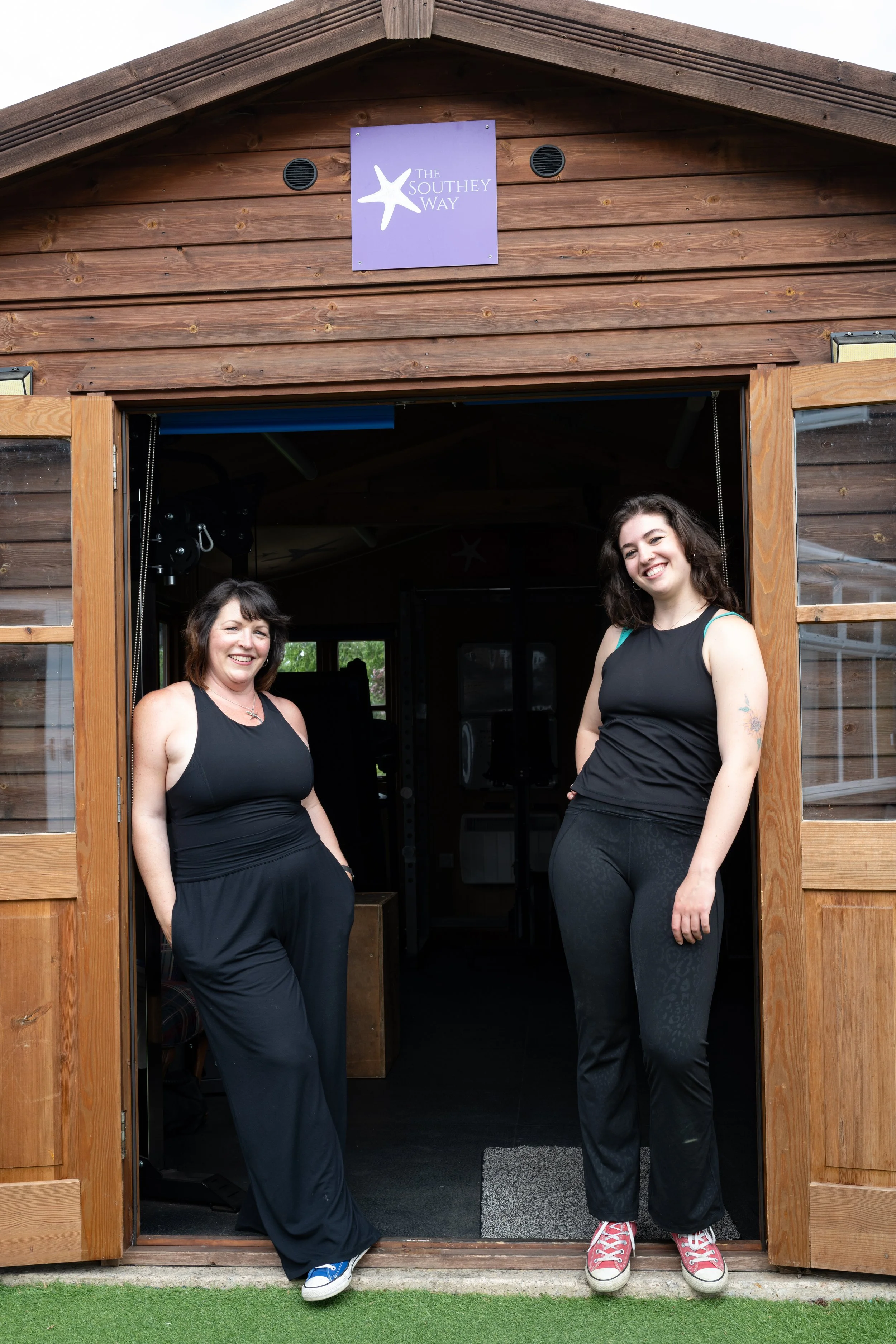 Co-directors Sara & Jenny standing in the doorway of a wooden structure labeled 'The Southey Way,' smiling at the camera.