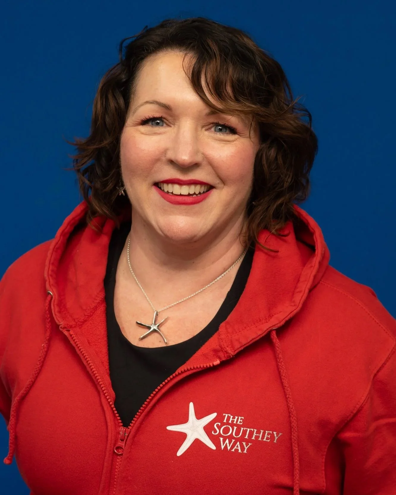 Sara Southey founder and co-director of the southey way, a woman with short, curly brown hair is wearing a red jacket with 'The Southey Way' logo, smiling against a blue background.