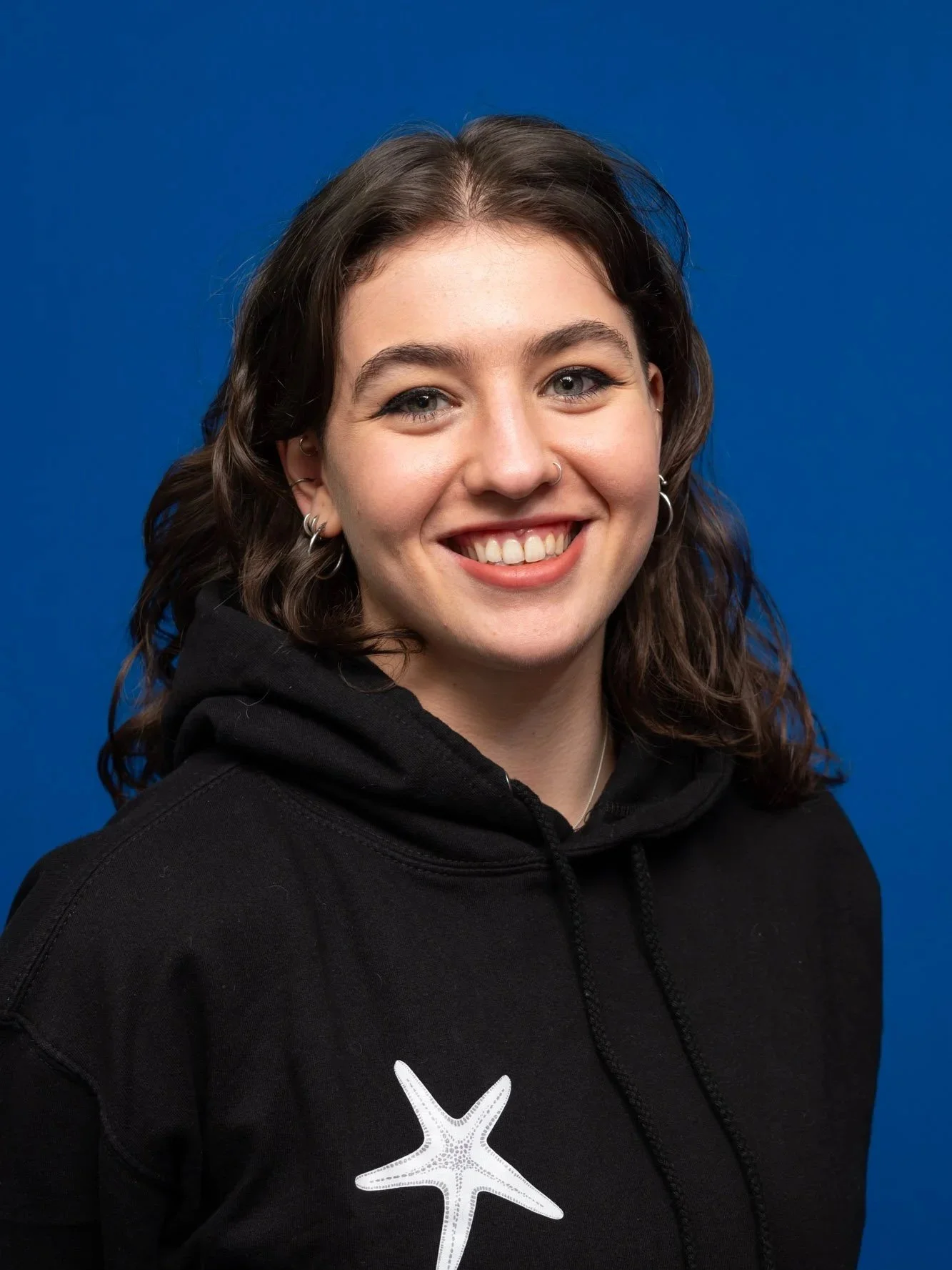 Jenny Southey co-director of the southey way, a woman with dark wavy hair smiling, wearing a black hoodie with 'The Southey Way' logo, earrings, and a nose ring, standing against a blue background.