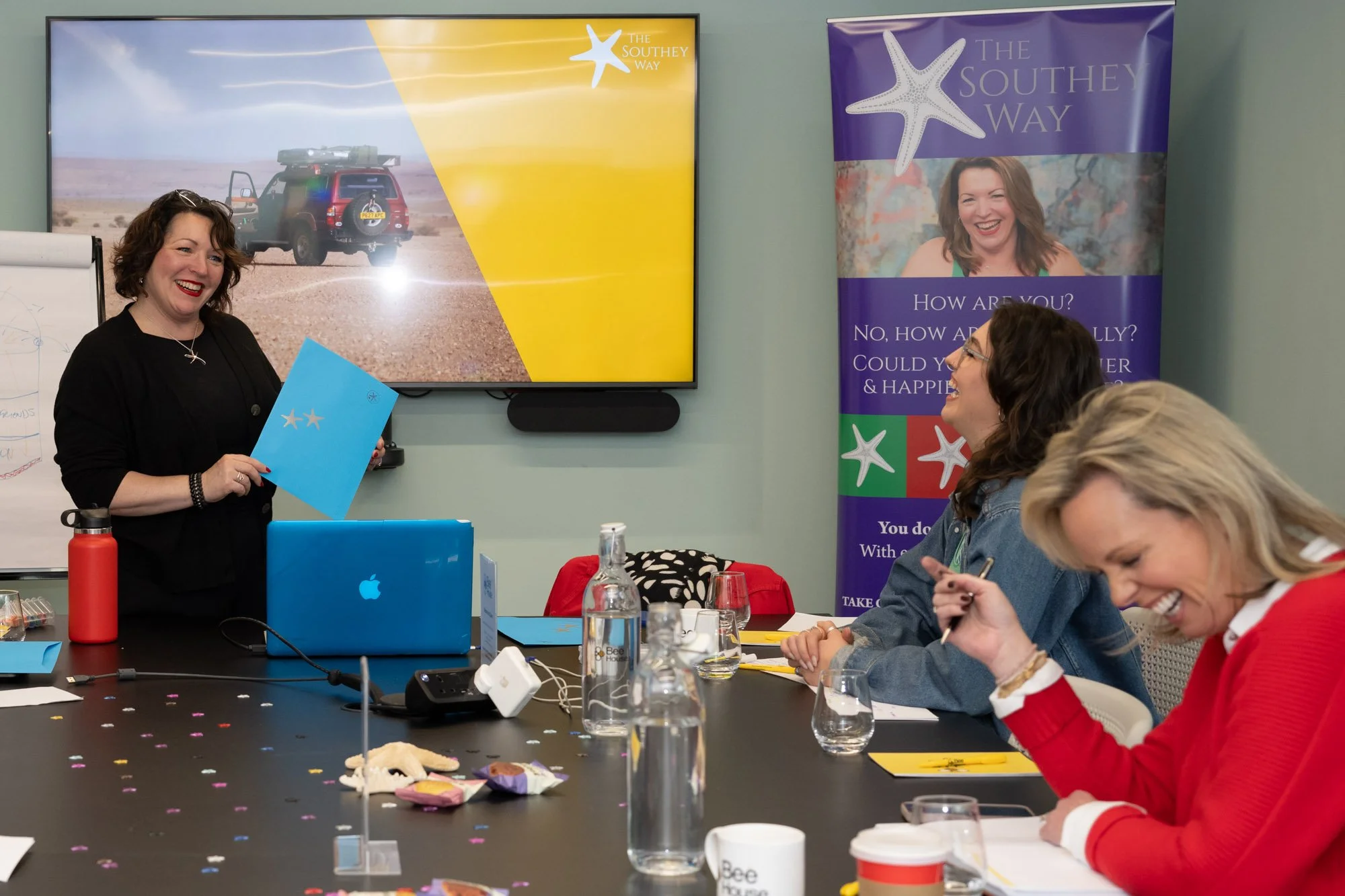 Sara's standing and smiling while holding a blue paper with two star cutouts, in a meeting room. Three women sit at a table, smiling and engaged. There are drinks, snacks, and confetti on the table, with a large screen and a banner in the background.