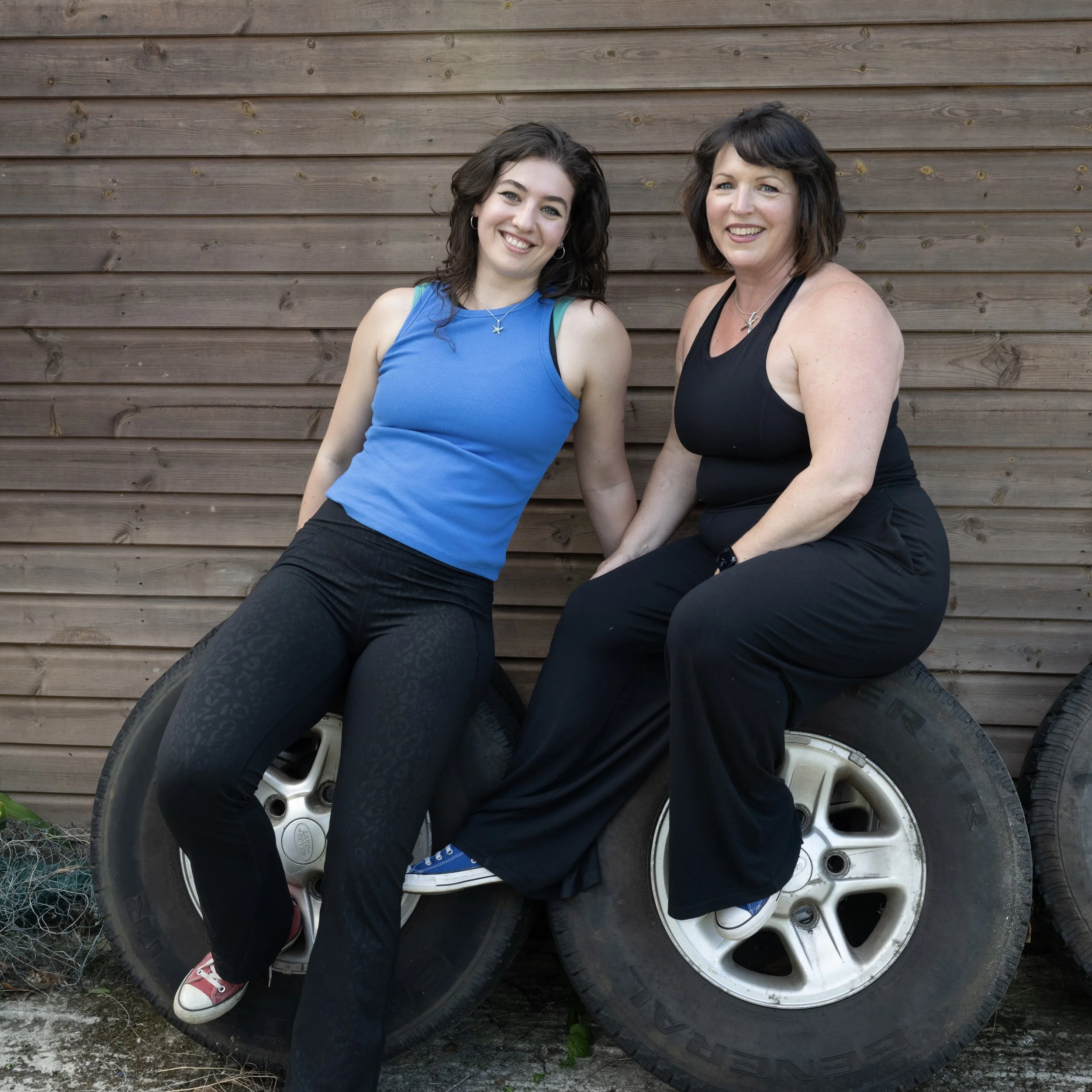 Co-directors Jenny & Sara sitting on tires in front of a wooden wall, smiling at the camera, one in a blue tank top and black pants, the other in a black tank top and black pants.