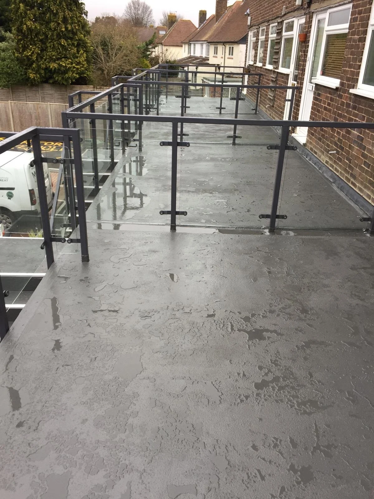 A concrete balcony with glass and metal railings attached to a brick building. Wet surface with water puddles, overlooking a residential neighborhood with trees and houses in the background.