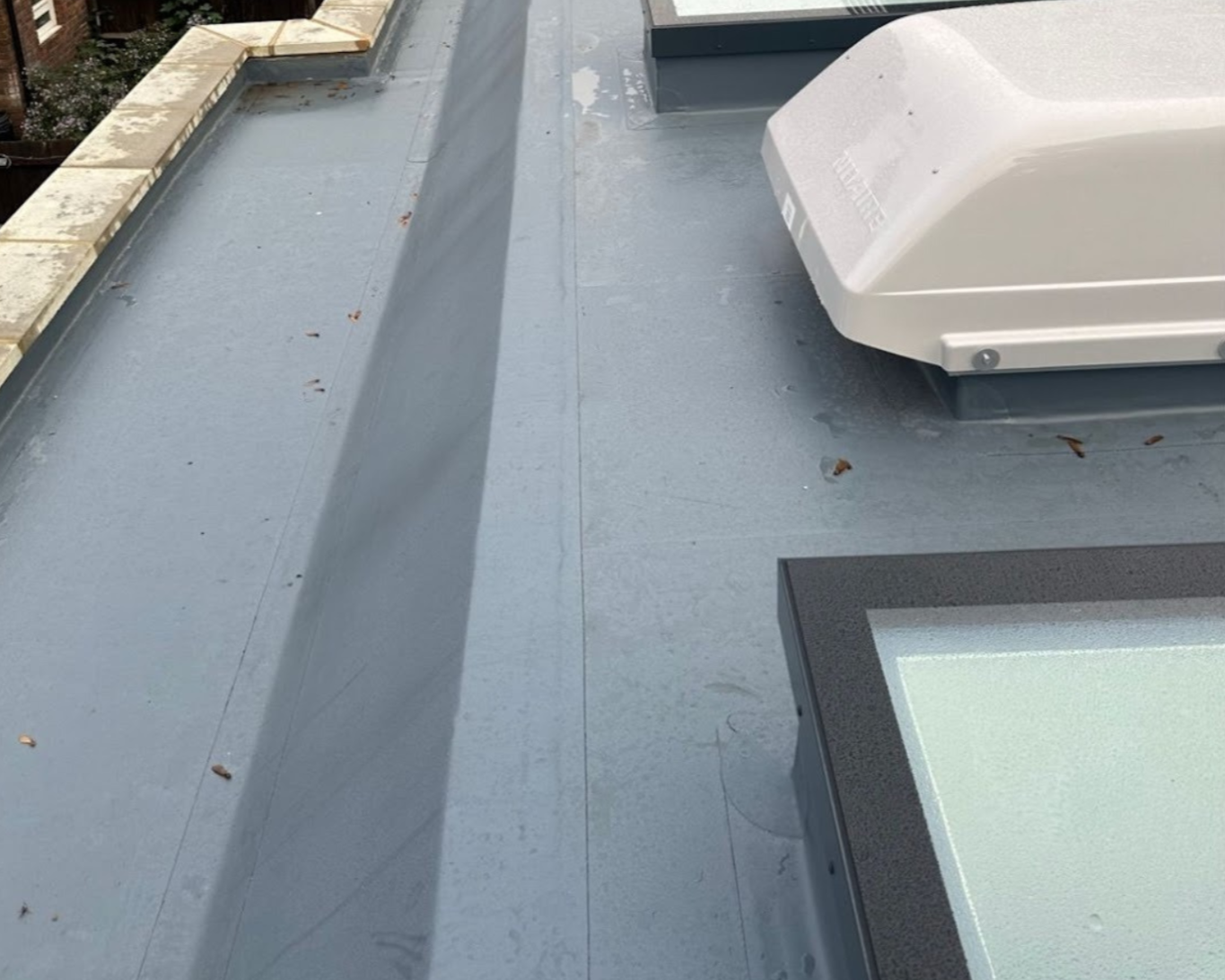 View of a flat rooftop with a gray surface, some scattered leaves, a white rooftop HVAC unit on the right, and a section of a window at the bottom right corner.