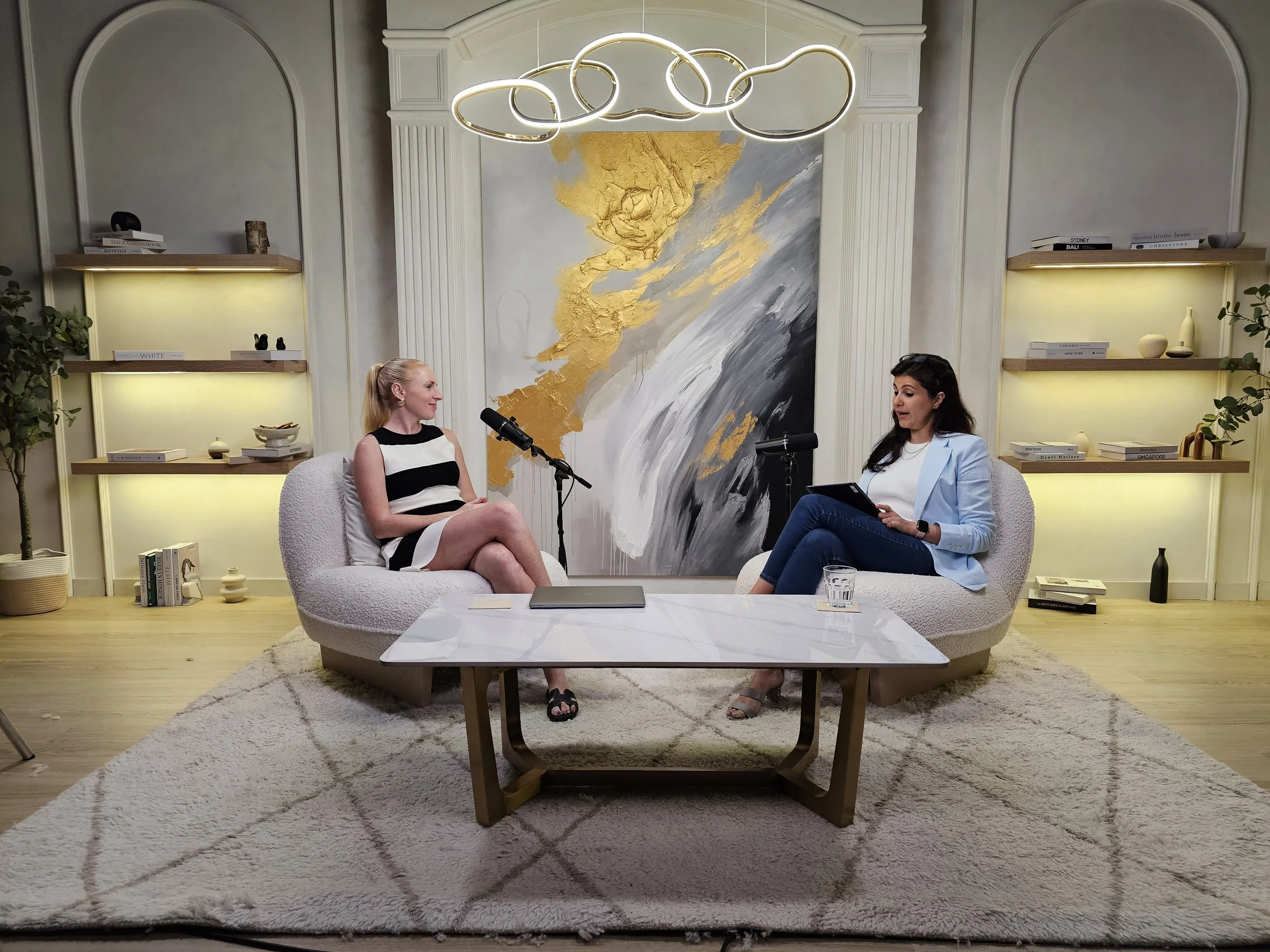 Two women are sitting in a modern, well-decorated living room, engaged in a conversation. Behind them is a large abstract painting with gold, gray, and white colors. The room features companies books on shelves, a white marble coffee table, and a plush white rug. There is stylish lighting overhead and potted plants in the background.