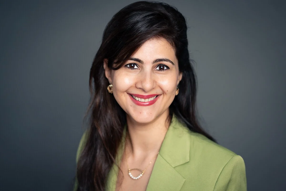 A professional woman with dark hair styled in loose waves, wearing a green blazer and gold jewelry, smiling against a grey background.