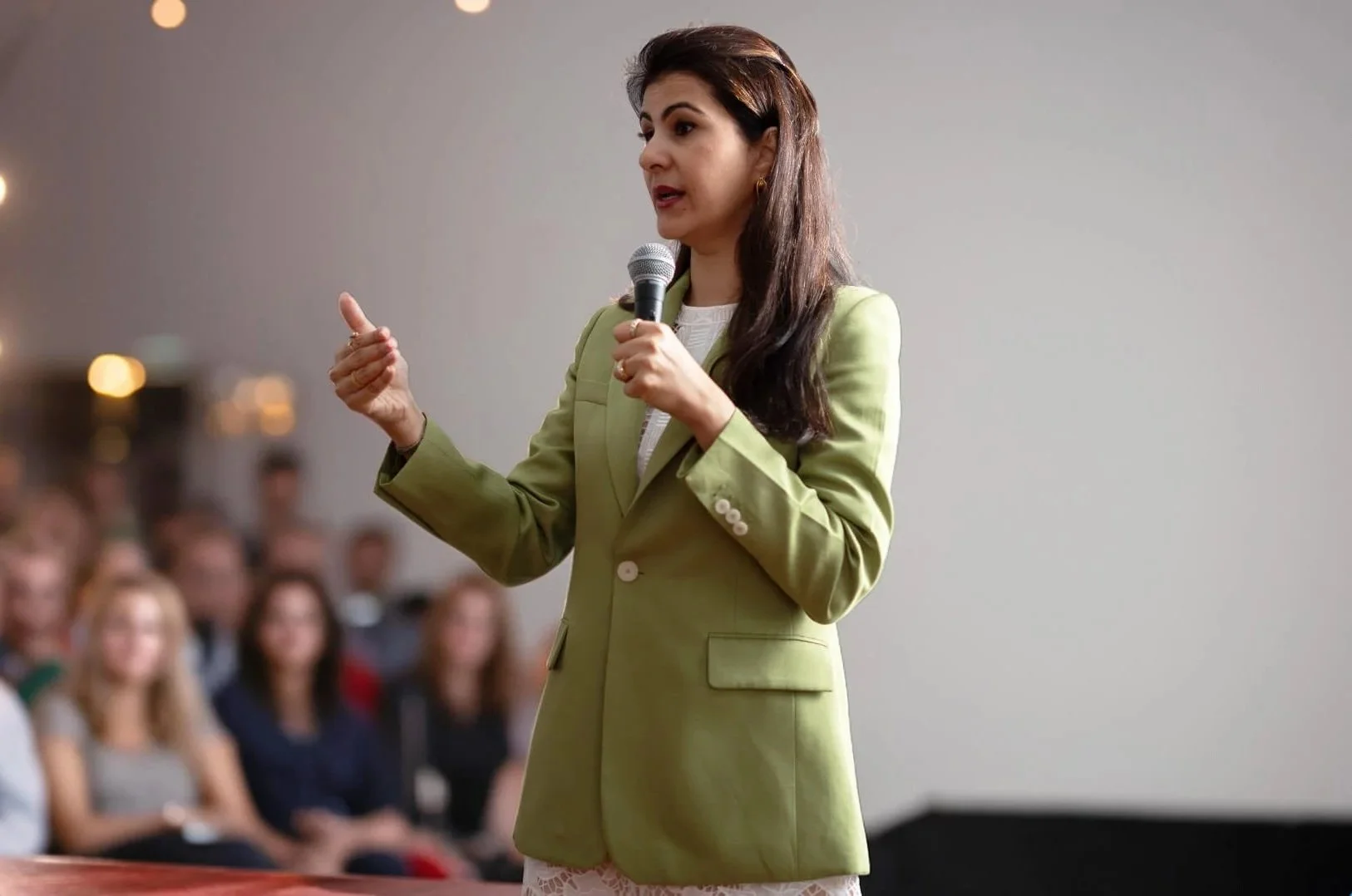 A woman in a light green blazer giving a presentation to an audience using a microphone.
