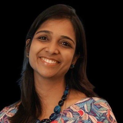 Close-up of a smiling woman with long dark hair, wearing a colorful patterned top and a blue beaded necklace, against a black background.