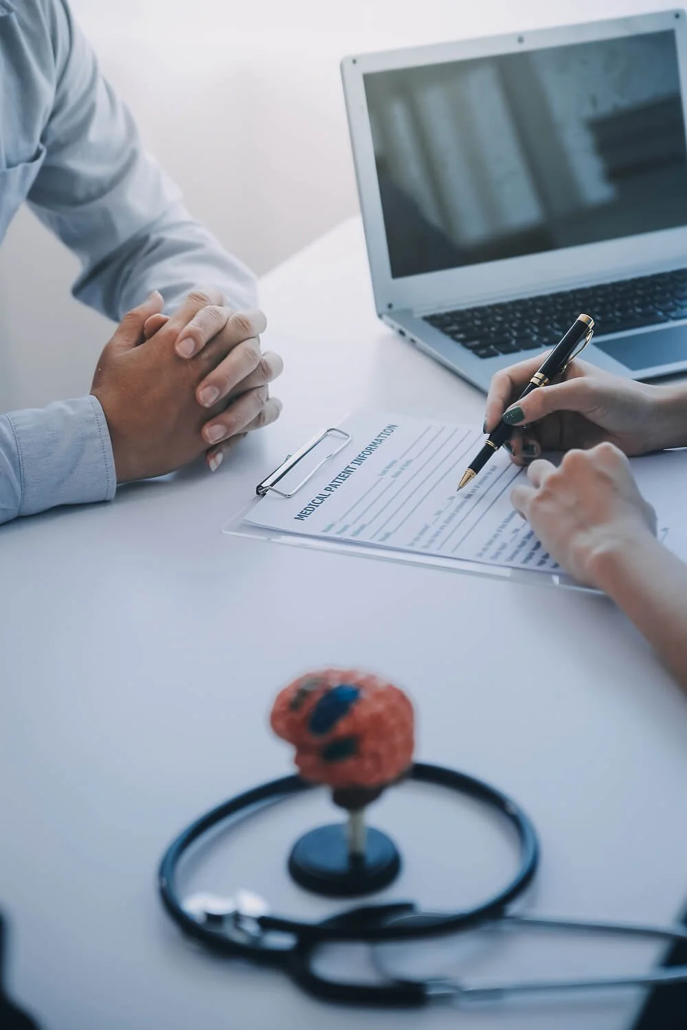 Doctor and patient with hands clasped during a medical appointment at a desk with a laptop and a medical patient information sheet. A stethoscope and a small model of a red blood cell are on the desk.