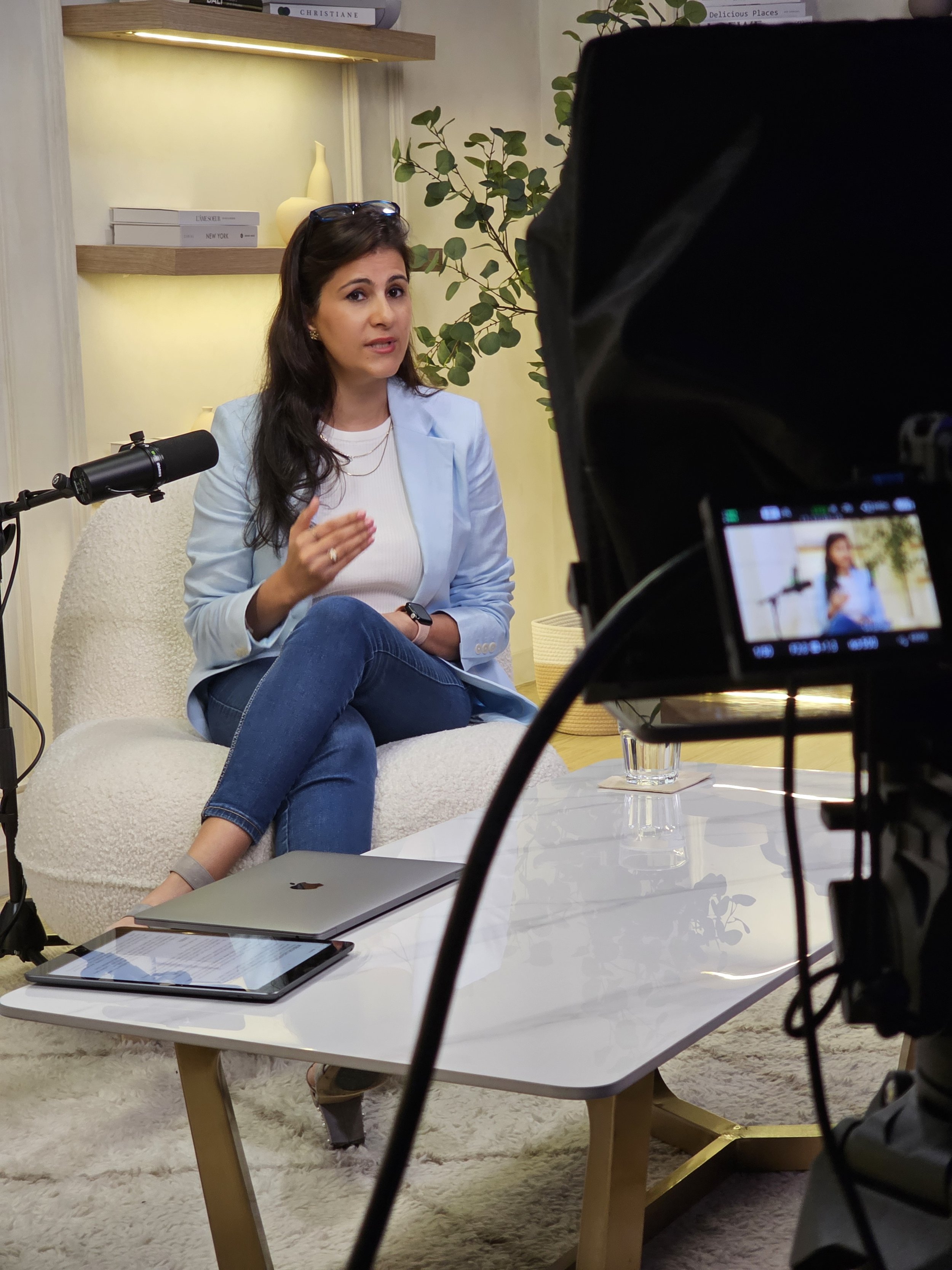A woman in a light blue blazer and white top is seated at a white coffee table, speaking during an interview or conversation, with a microphone placed nearby. A camera captures her from in front, showing her gesturing with her right hand. The background includes a bookshelf, a plant, and decorative vases, with a laptop and tablet on the table.
