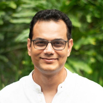 Portrait of a man with glasses smiling outdoors with green foliage in the background.
