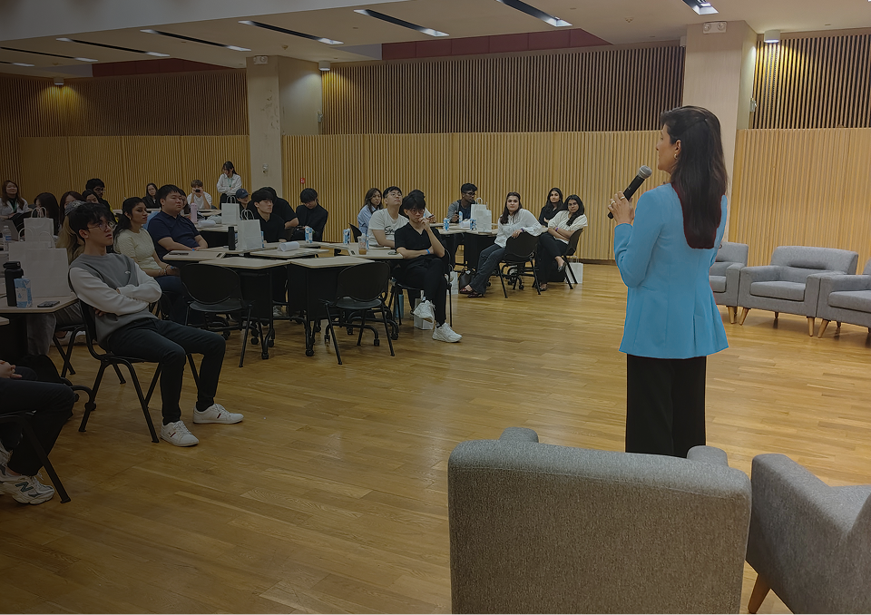 A woman giving a presentation with a microphone to a seated audience in a modern room with wooden paneling.