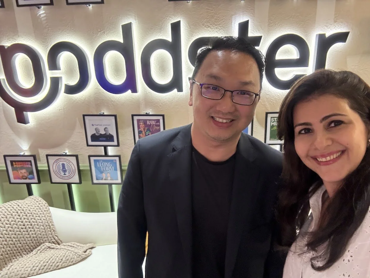 Two people smiling for a selfie in front of a wall with illuminated 'podcaster' sign and framed podcast icons.
