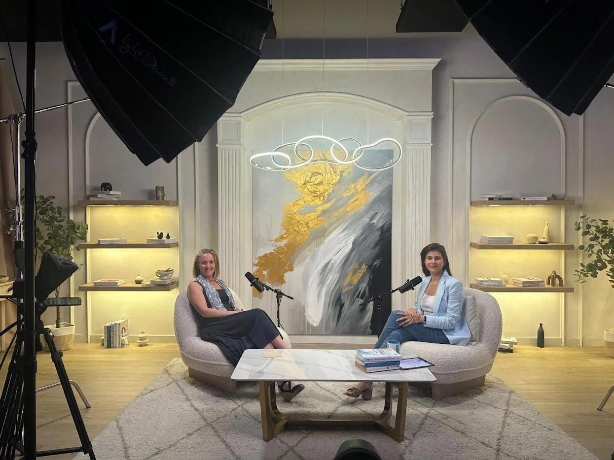 Two women sitting on cream-colored curved couches in a studio set, with a large abstract painting behind them, shelves with books and decor on either side, a white marble coffee table with books, and professional studio lighting.