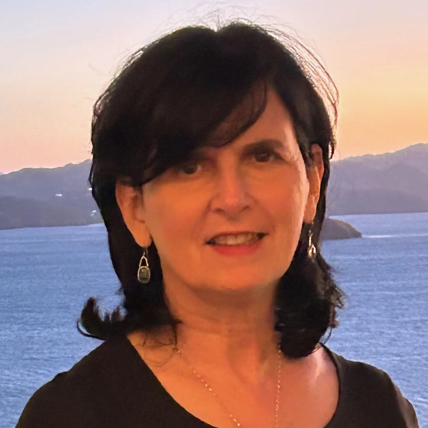 A woman with shoulder-length dark hair, wearing earrings, a necklace, and a black top, standing near a body of water during sunset or sunrise, with mountains in the background.