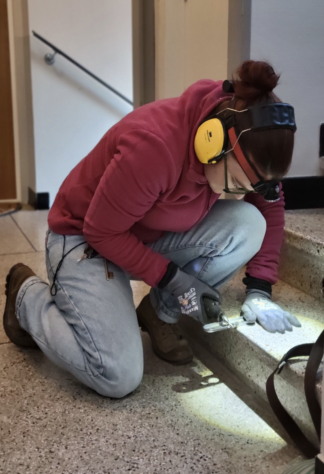 Une personne portant un casque antibruit, des gants et utilisant une perceuse électrique en position agenouillée, travaillant sur un rebord en béton.