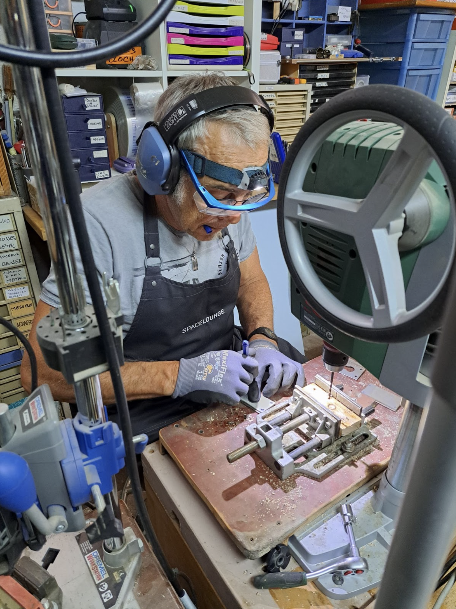Un homme porte des lunettes de protection, des écouteurs et un tablier, travaillant avec un microscope de précision sur une pièce en métal dans un atelier rempli d'outils et de rangements.