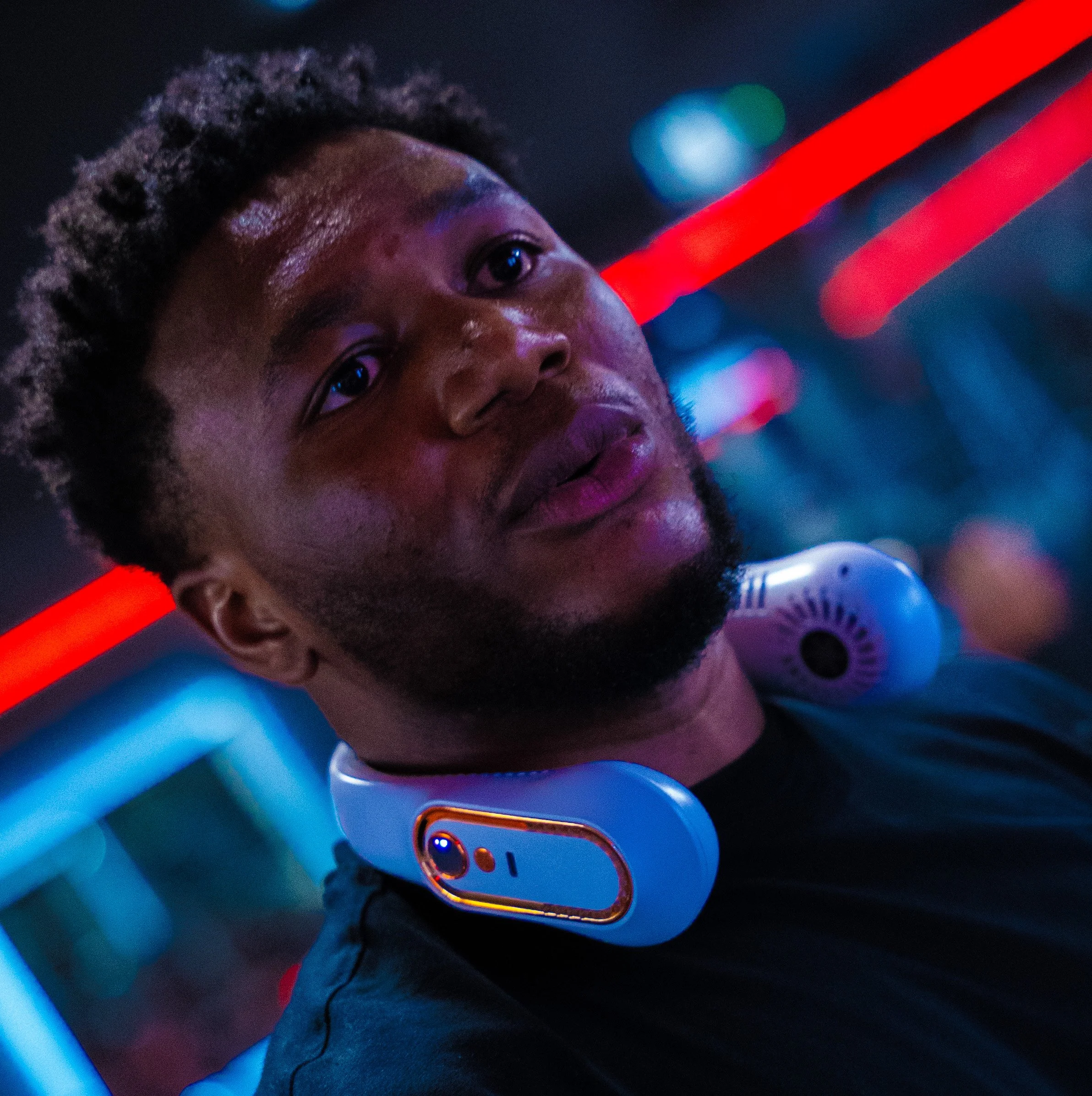A young man with dark skin and short curly hair, wearing a black shirt and a futuristic white choker-like device with a built-in camera, is in a neon-lit environment with red and blue lights.