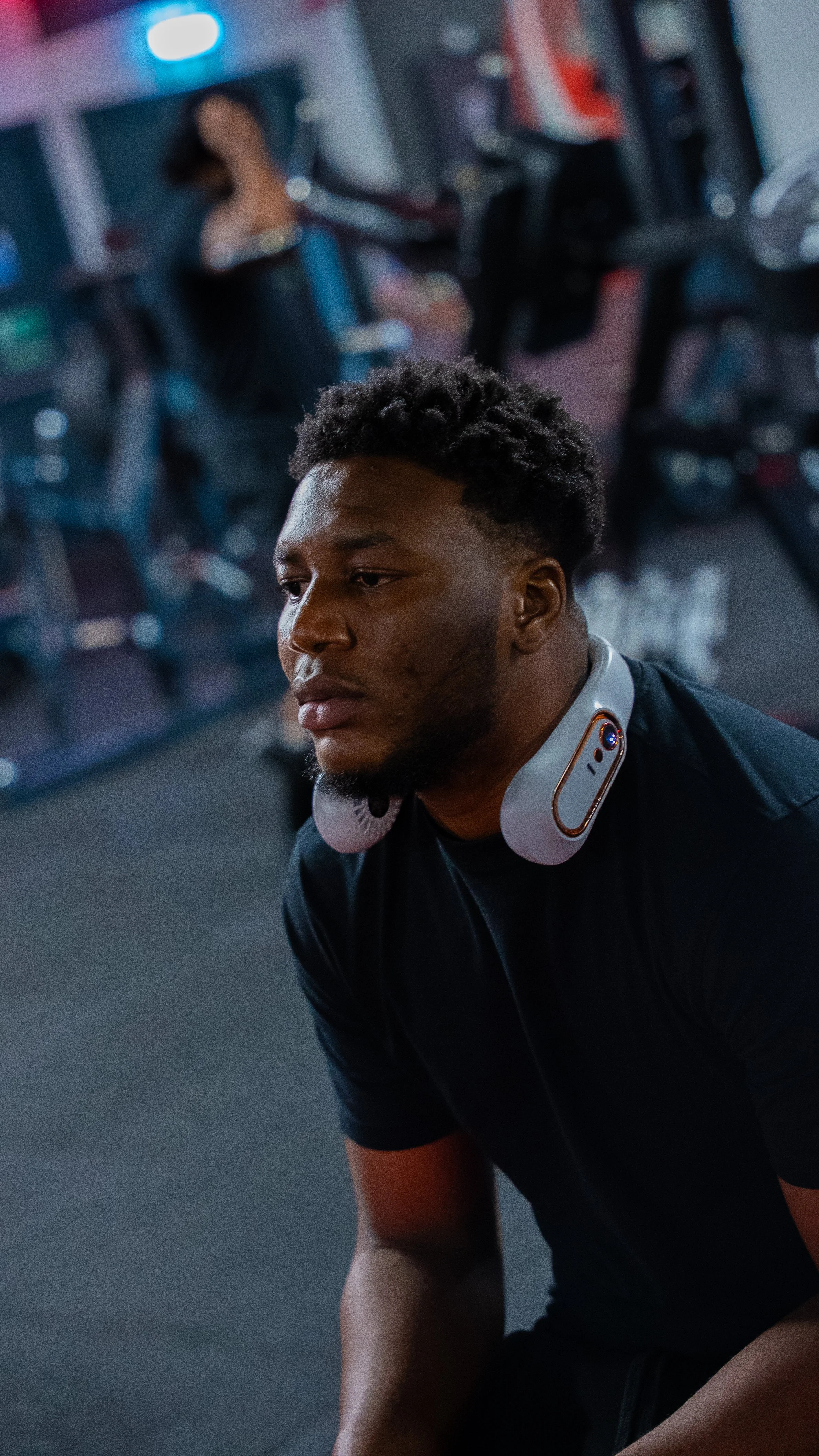 A young man with a beard and short, curly hair sitting on the gym floor, wearing headphones around his neck, with gym equipment and a woman exercising in the background.