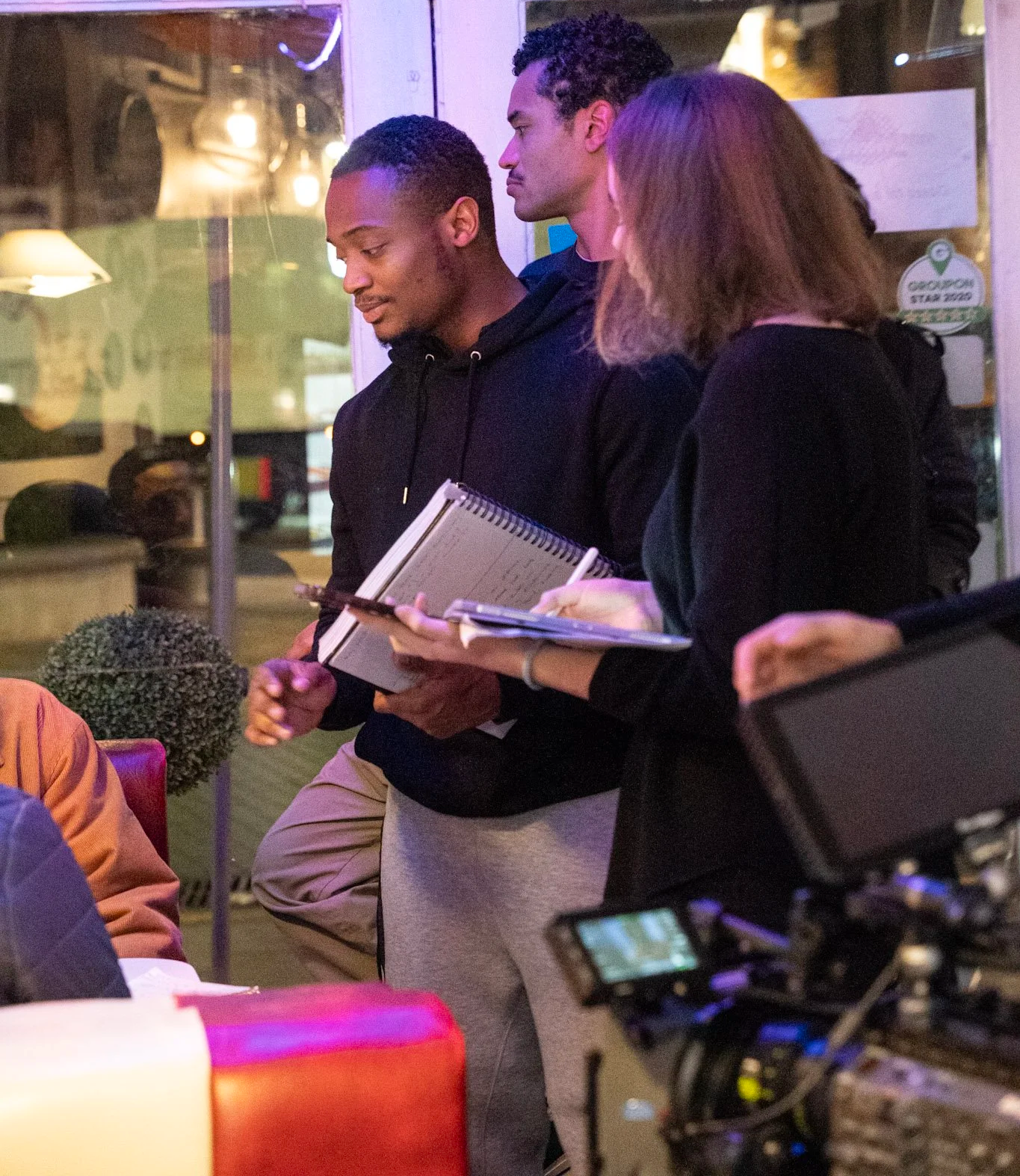 Three people standing indoors, engaged in a discussion, with one person holding a notepad and a phone, and a camera in the foreground.