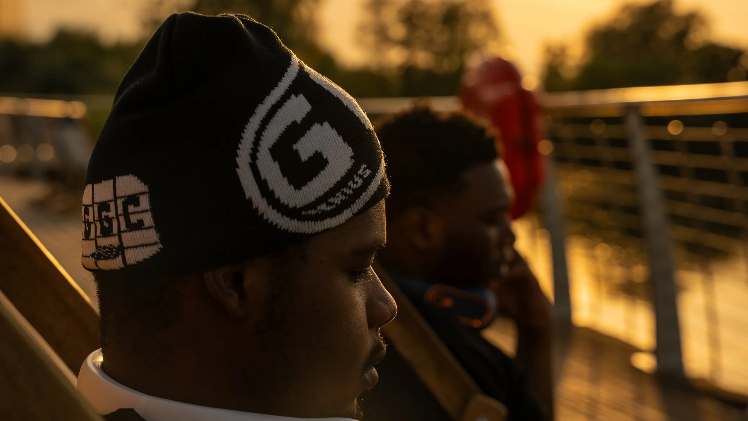 Two young men sitting on a bench during sunset, one wearing a black beanie with white patch and the other talking on a mobile phone.