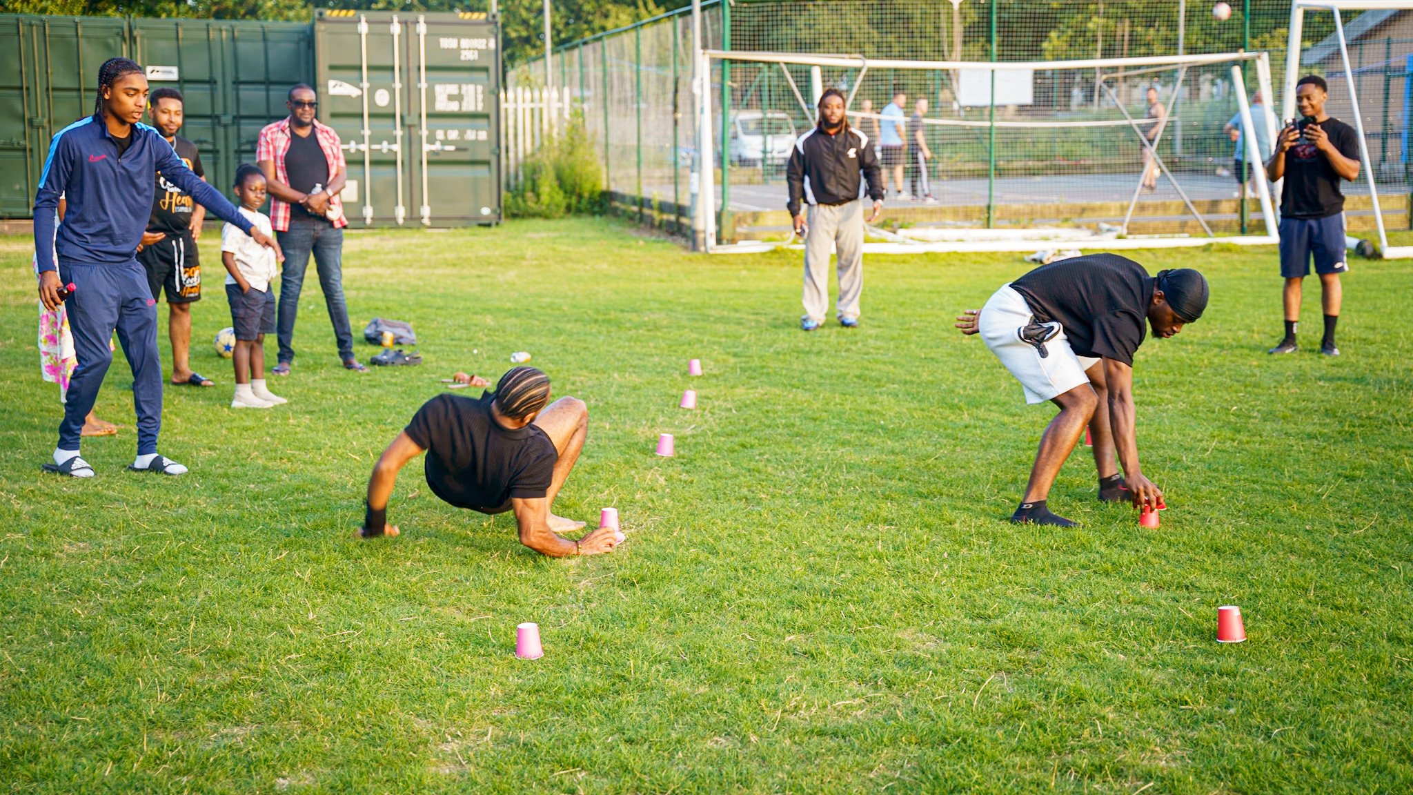 People participating in an outdoor fitness game involving crawling over pink cups on a grassy field, with spectators watching.