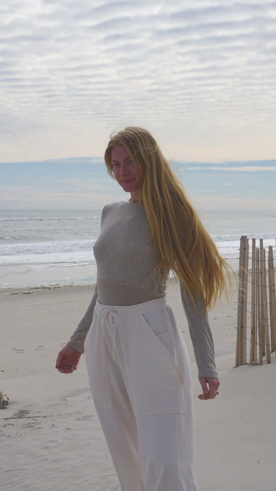 A woman with long blonde hair wearing a gray long-sleeve shirt and white drawstring pants standing on a sandy beach near the ocean with cloudy sky overhead.