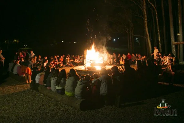 Pessoas reunidas ao redor de uma fogueira durante a noite, em um ambiente ao ar livre ao redor de árvores.