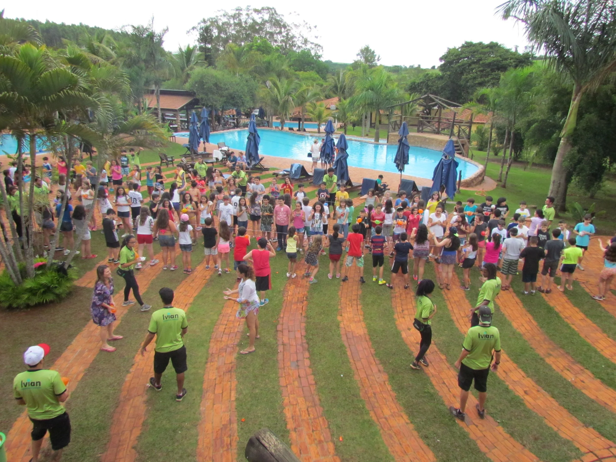 Muitas pessoas participando de uma atividade ao ar livre próximo a uma piscina, rodeadas de árvores e vegetação, com guarda-sóis e cadeiras ao redor.