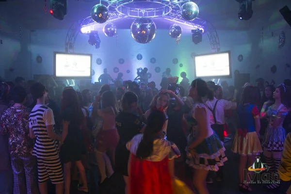 Pessoas dançando em uma festa com iluminação colorida, espelhos decorativos e telões ao fundo.
