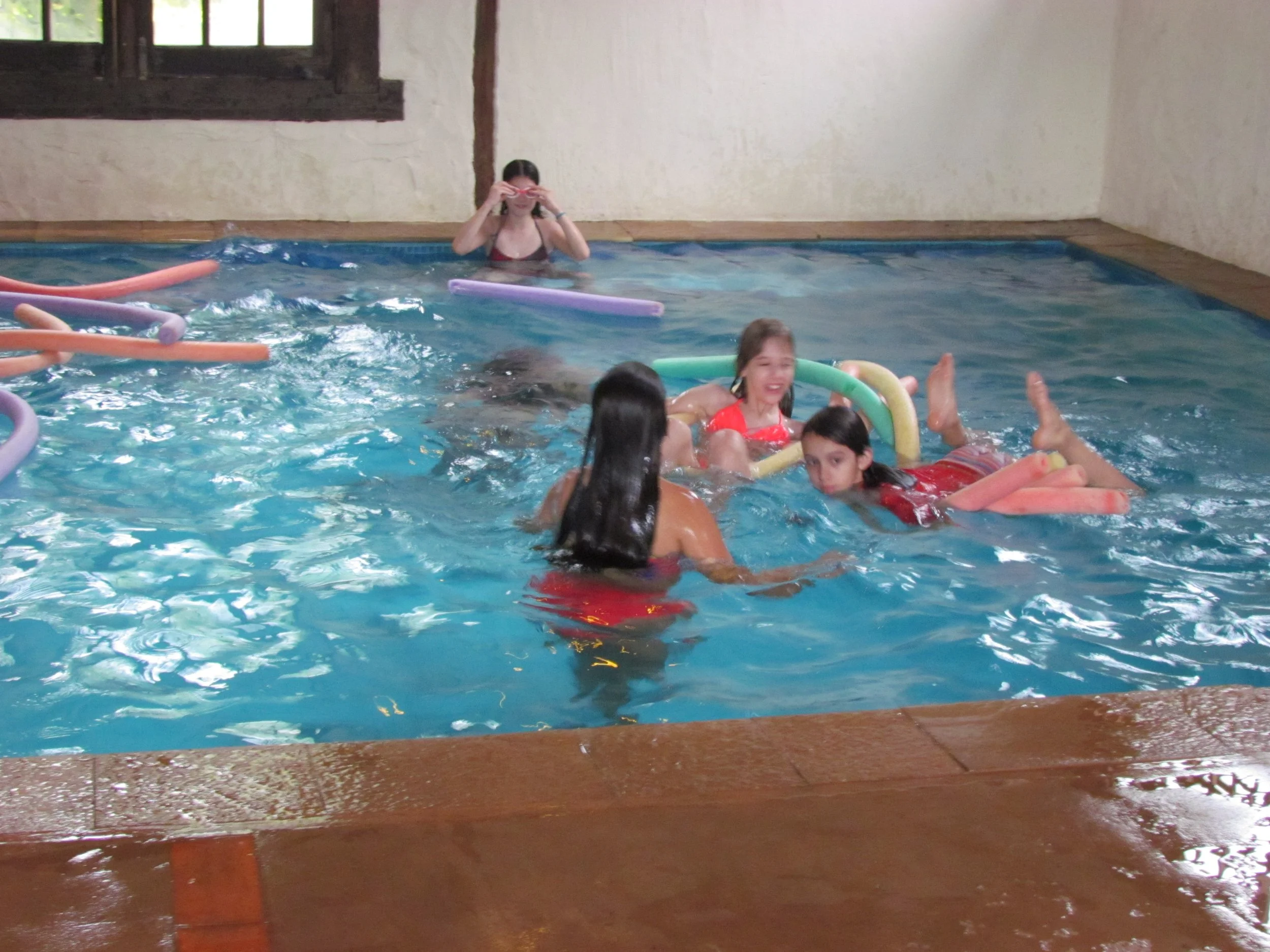 Crianças brincando na piscina com bóias de brinquedo, uma mulher assistindo e ajustando seus óculos na piscina. Algas e janelas de madeira ao fundo.