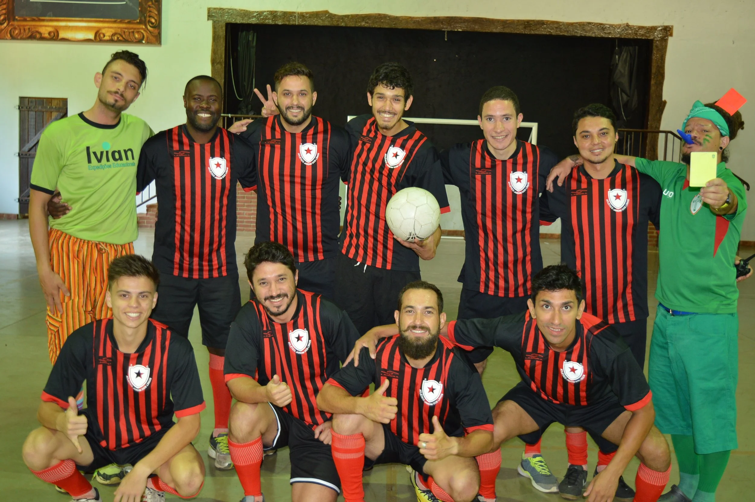 Grupo de pessoas posando com uniforme de futebol, alguns segurando uma bola, em ambiente interno, todos sorrindo e fazendo gestos de apoio.