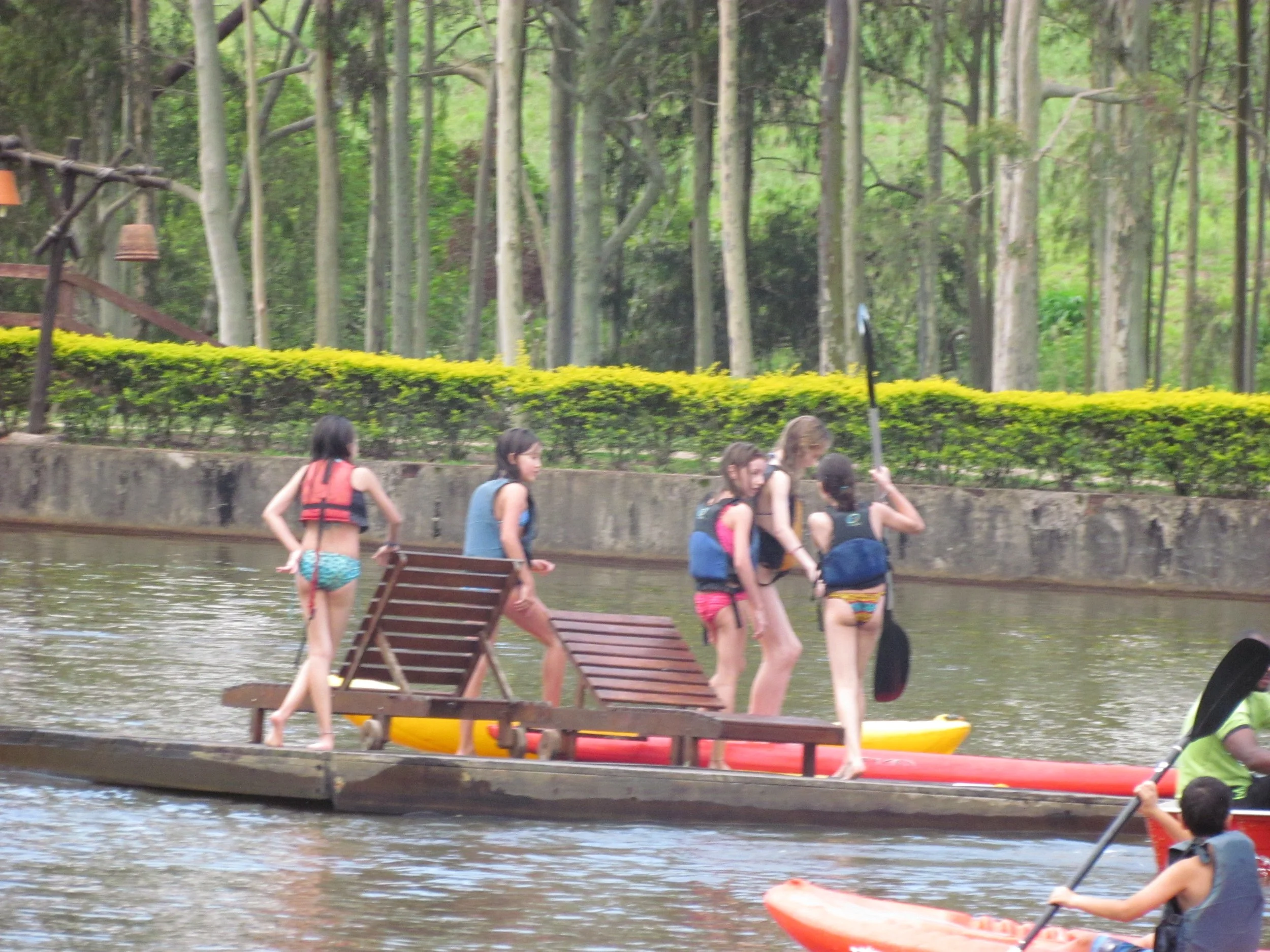 Grupo de pessoas jovens com coletes salva-vidas, saindo de um barco de vários caiaques na água, na floresta. Algumas pessoas estão usando chapéus e há vegetação densa ao fundo.