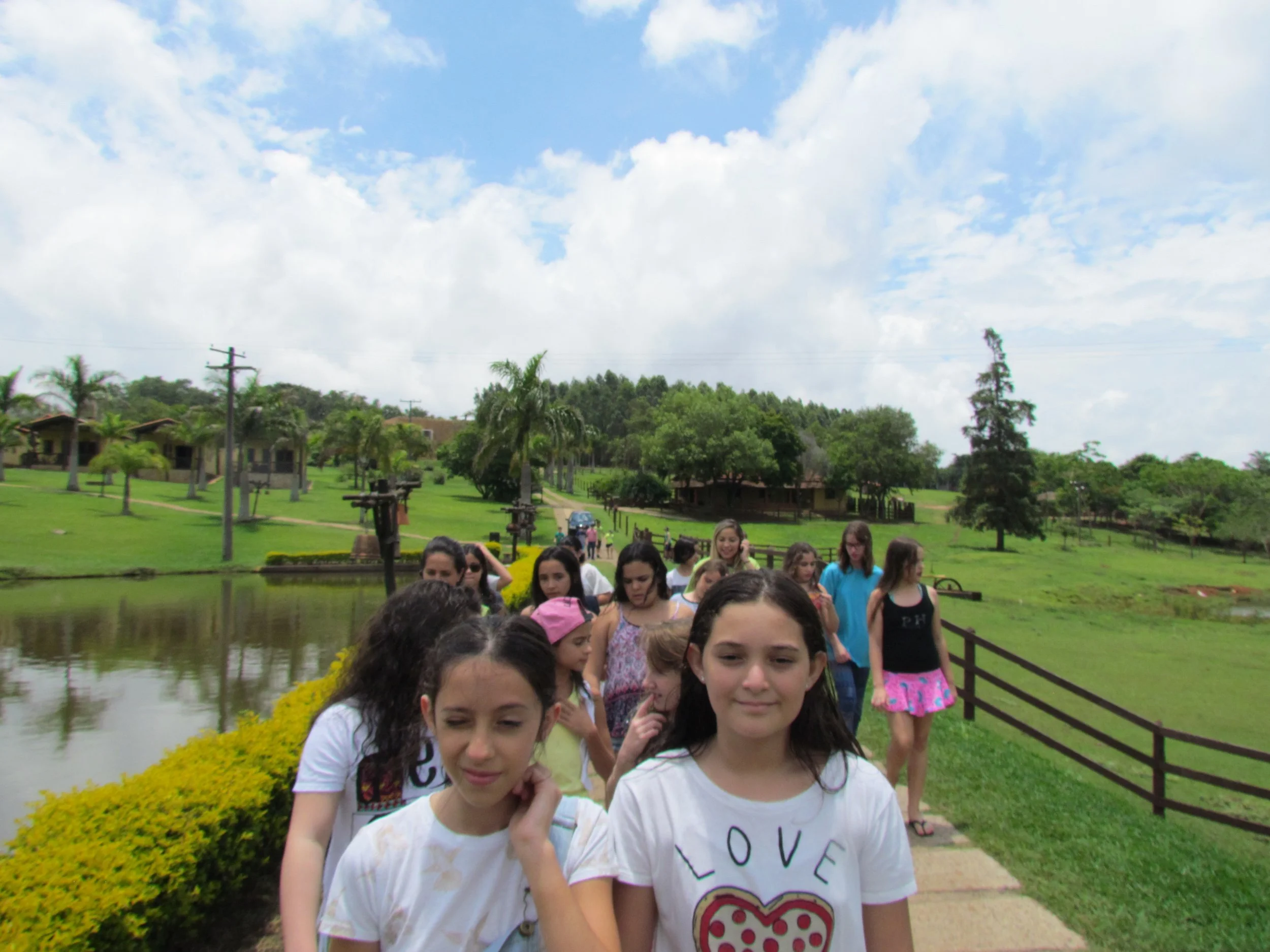 Grupo de jovens caminhando por uma ponte de madeira sob um cenário de árvores, lagos e céu com nuvens.