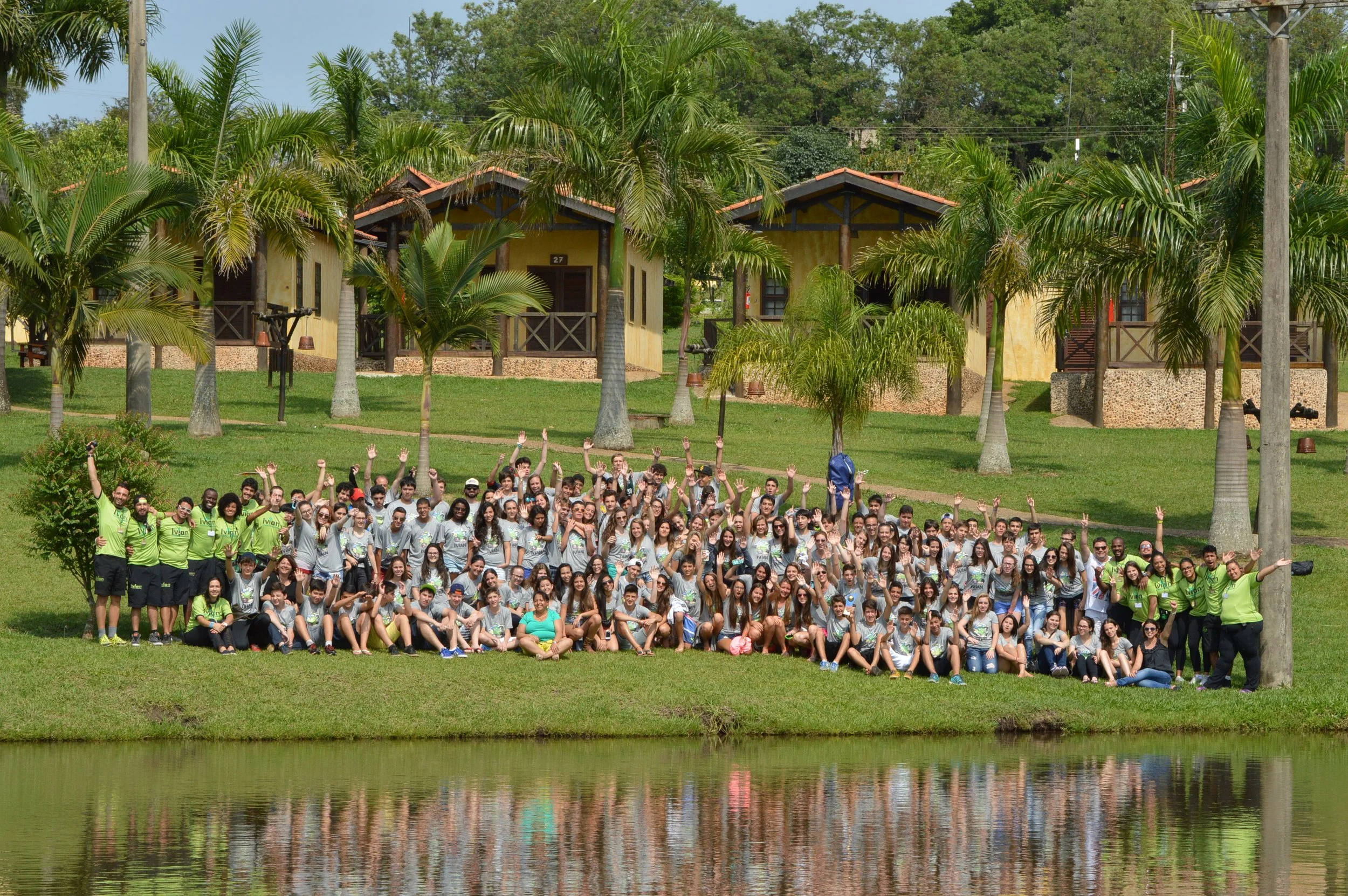 Grupo grande de pessoas, incluindo jovens e adultos, posando na frente de uma casa colorida com paredes amarelas e telhados vermelhos, rodeada por árvores e palmeiras altas, ao lado de um lago com reflexo na água.