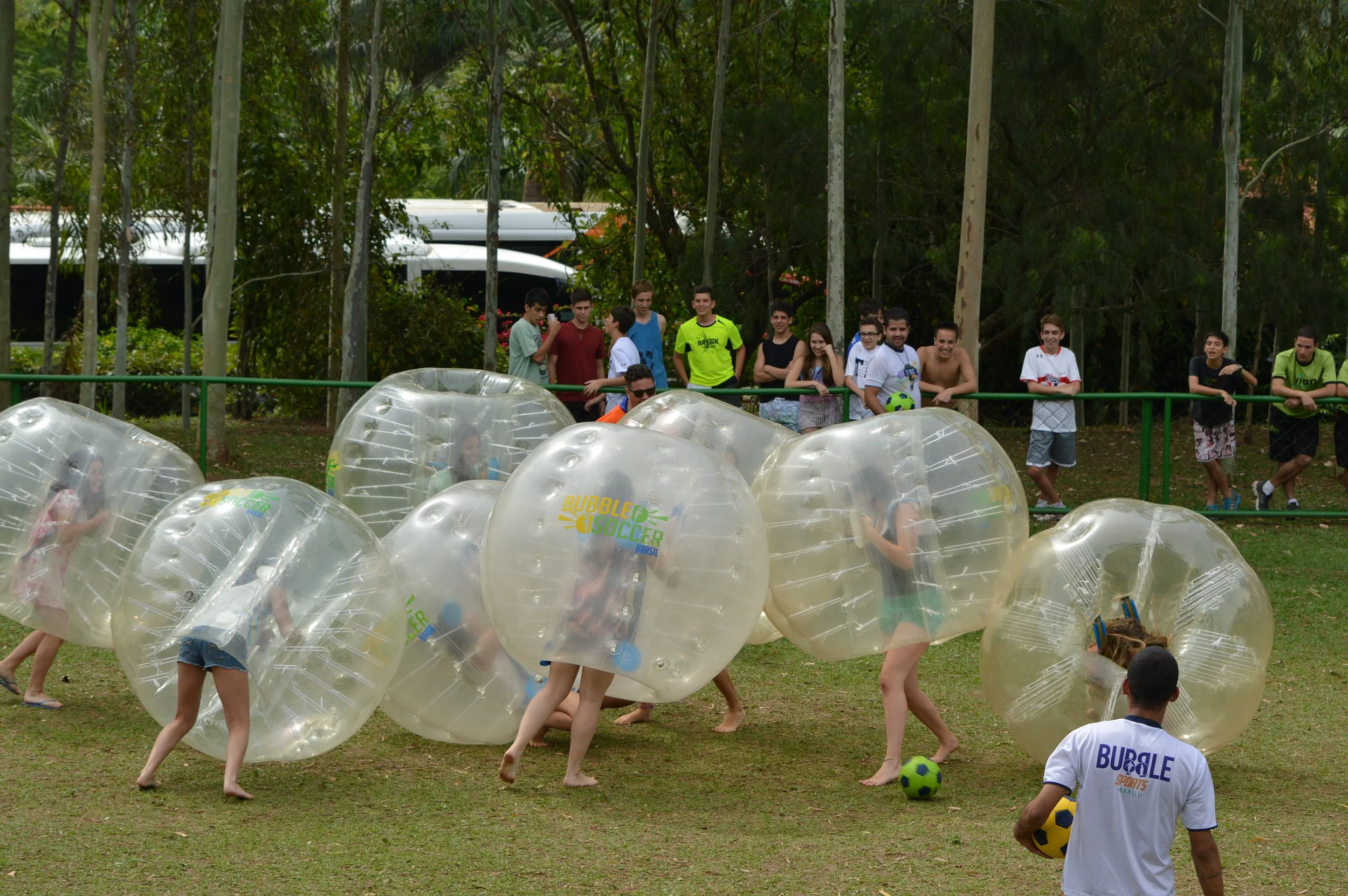 Grupo de crianças brincando com bolas de futebol e bolhas de plástico no parque, algumas crianças estão deitadas e outras em pé, com árvores ao fundo.