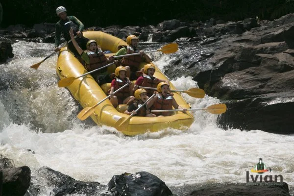 Grupo de pessoas em um barco amarelo de rafting descendo uma corredeira de rio com águas brancas, usando capacetes e coletes salva-vidas, ao redor de rochas e vegetação.