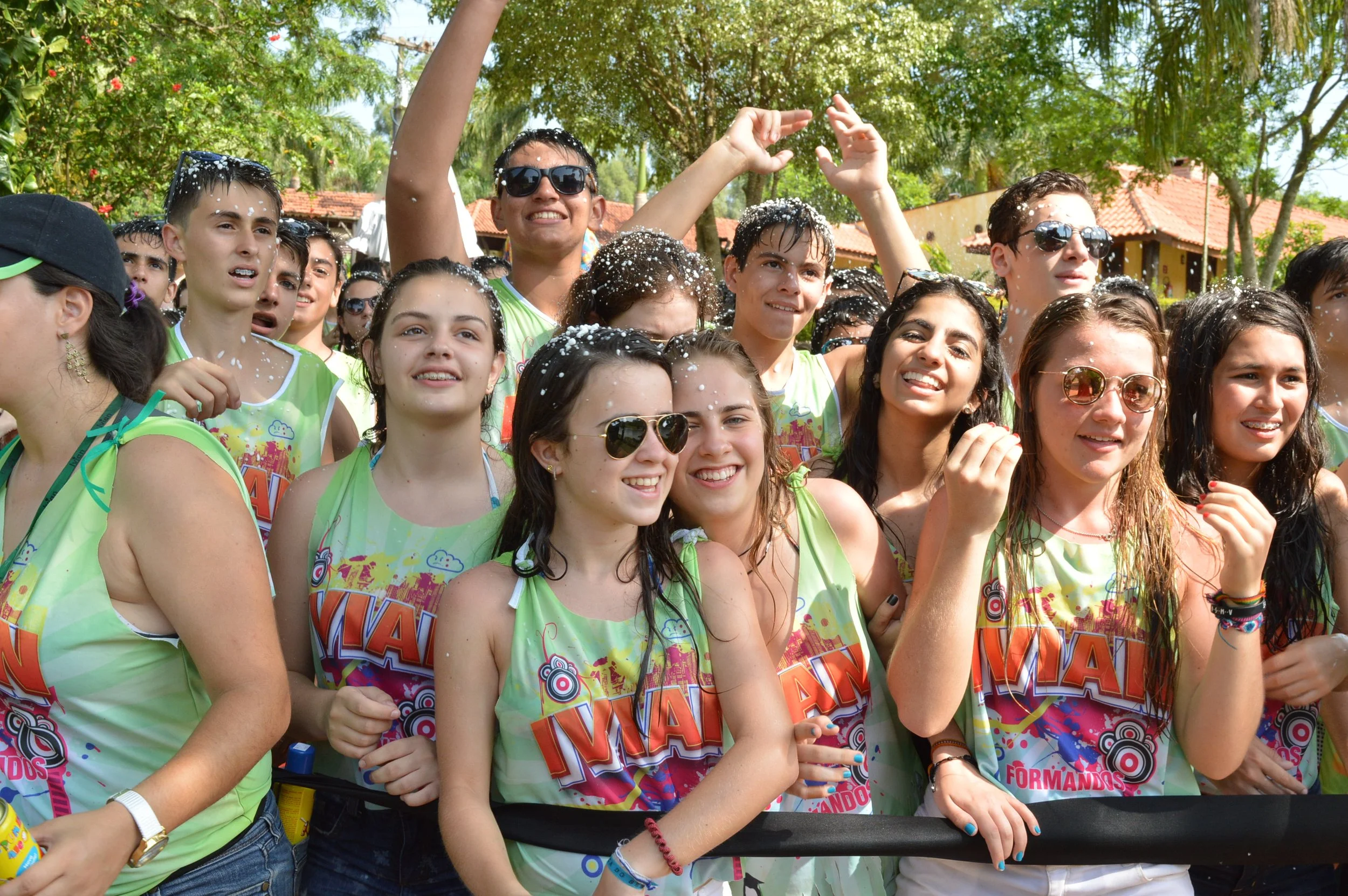 Grupo de jovens sorrindo e celebrando ao ar livre com roupas coloridas em um evento, com árvores ao fundo e pessoas usando óculos de sol.