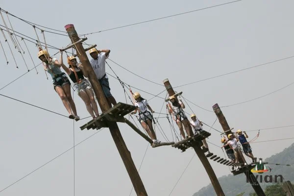 Pessoas atravessando uma ponte de corda suspensa em uma linha de energia elétrica, usando chapéus e roupas casuais, com fundo de céu nublado e uma paisagem distante.