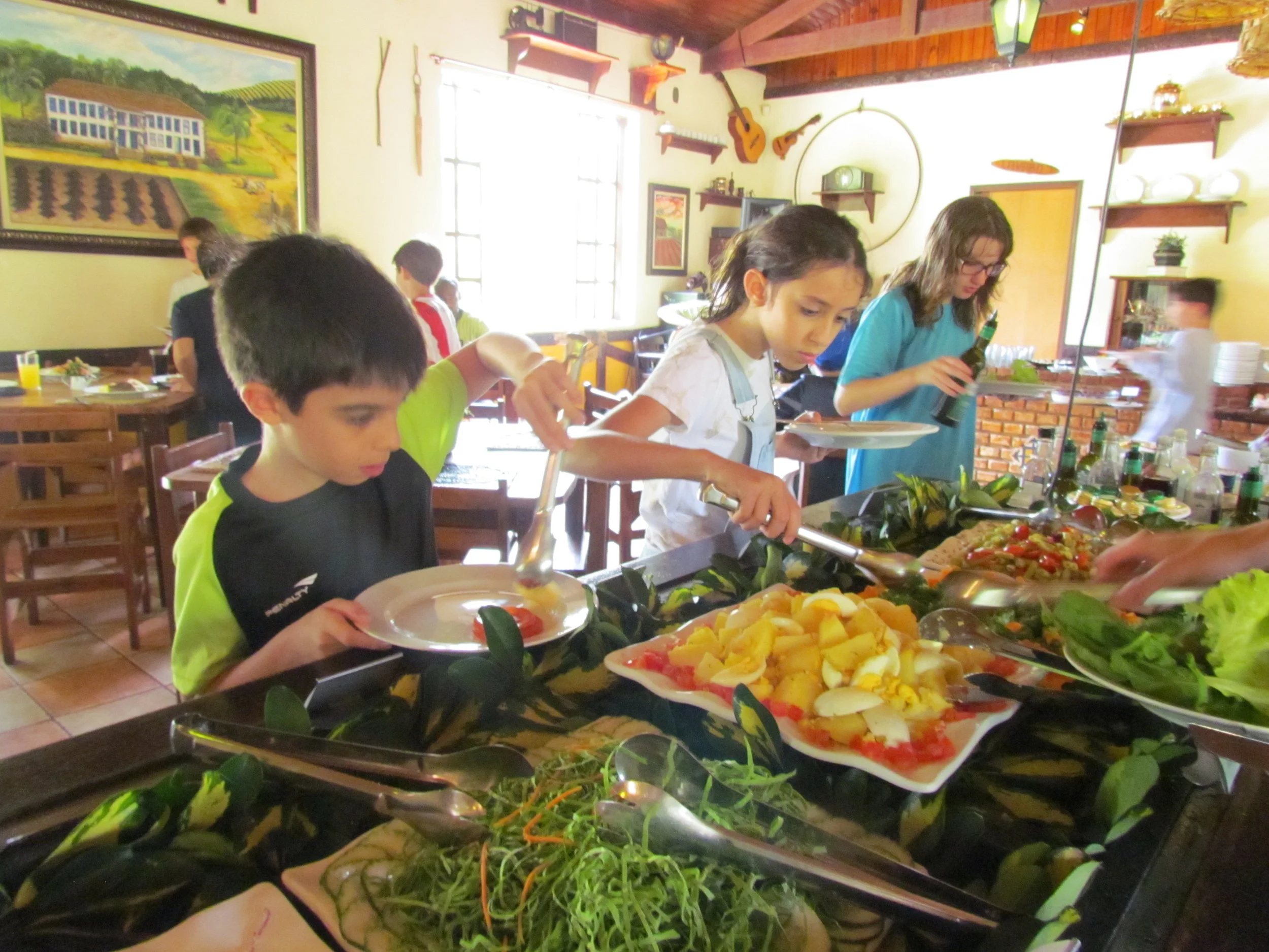 Pessoas em uma sala de jantar se servindo de um buffet com várias opções de alimentos, incluindo saladas, frutas e pratos diversos.