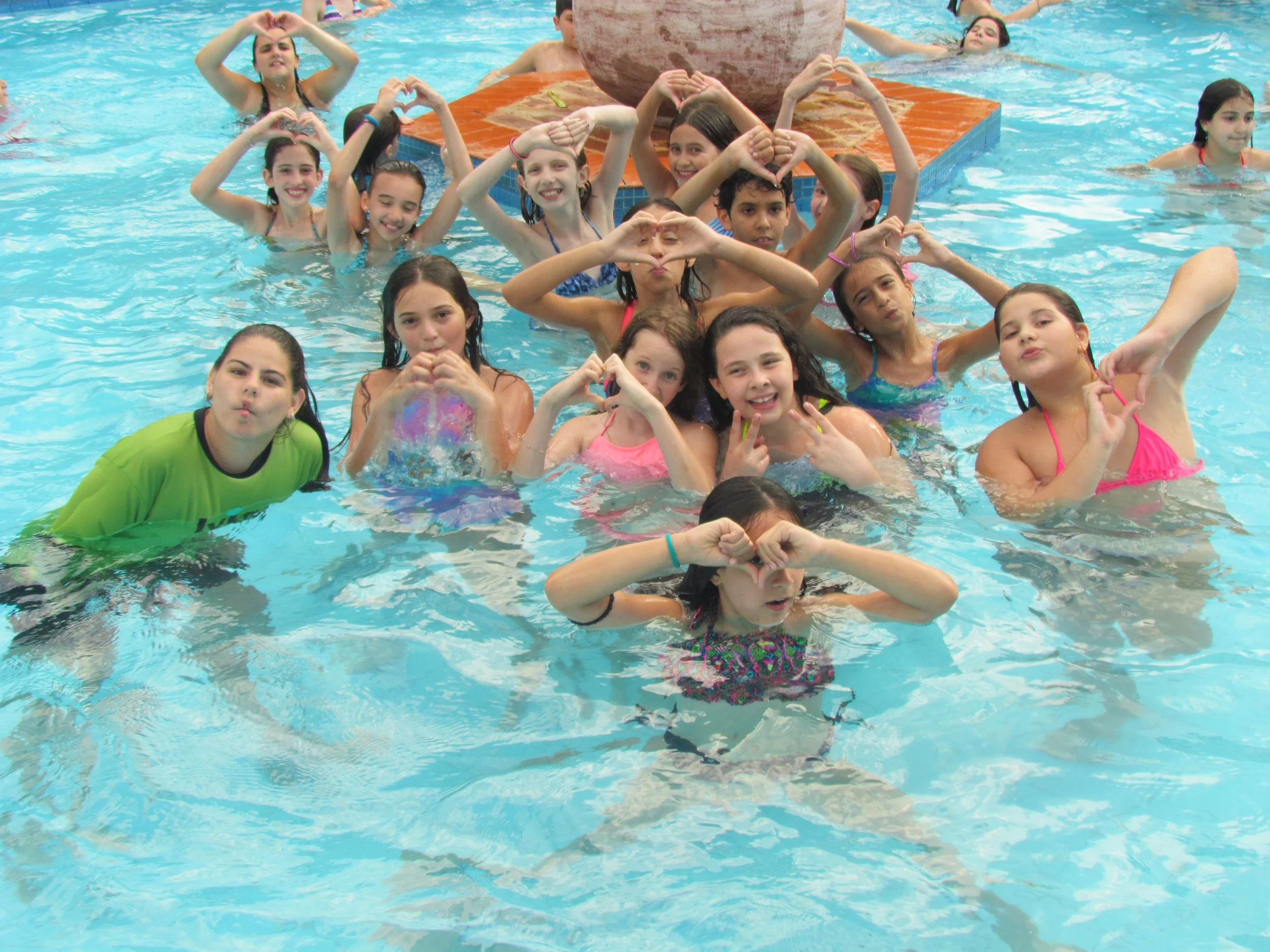 Grupo de crianças nadando na piscina, fazendo formas com as mãos e sorrindo.