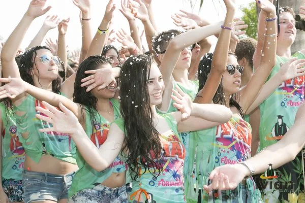 Grupo de jovens comemorando em festa ao ar livre, usando camisetas coloridas com tema de formatura, algumas com óculos de sol, com expressões felizes e braços levantados.